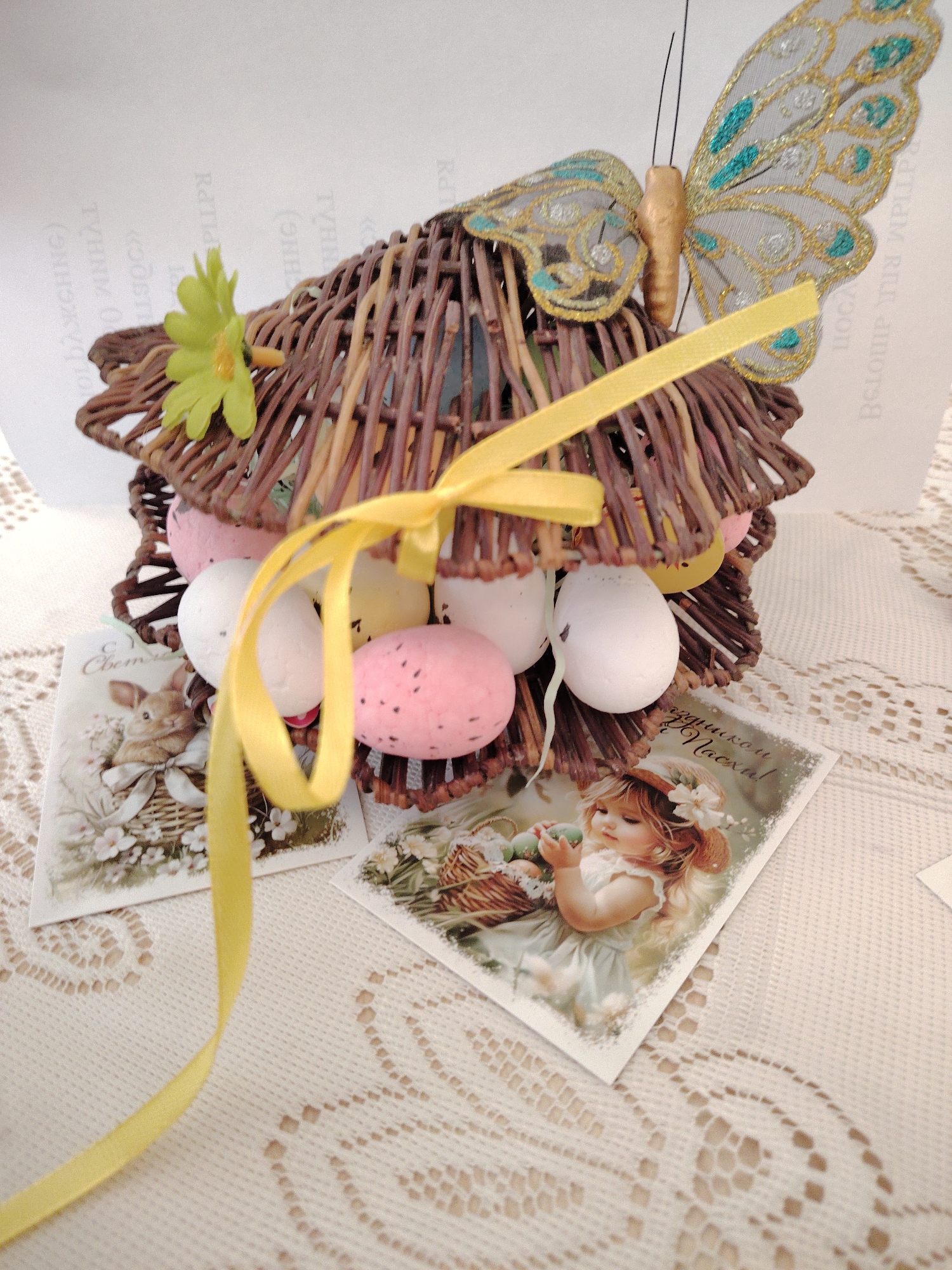 The image shows a decorative Easter basket. The basket is made of woven twigs and is decorated with a yellow ribbon, a green flower, and a butterfly. The basket contains several Easter eggs, some white and some pink with dark spots. Two postcards are in front of the basket, one with a rabbit and the other with a young girl holding a basket. The scene is set on a lacy tablecloth. The background is out of focus. It is unknown if this image was taken in Sarapul, Russia.