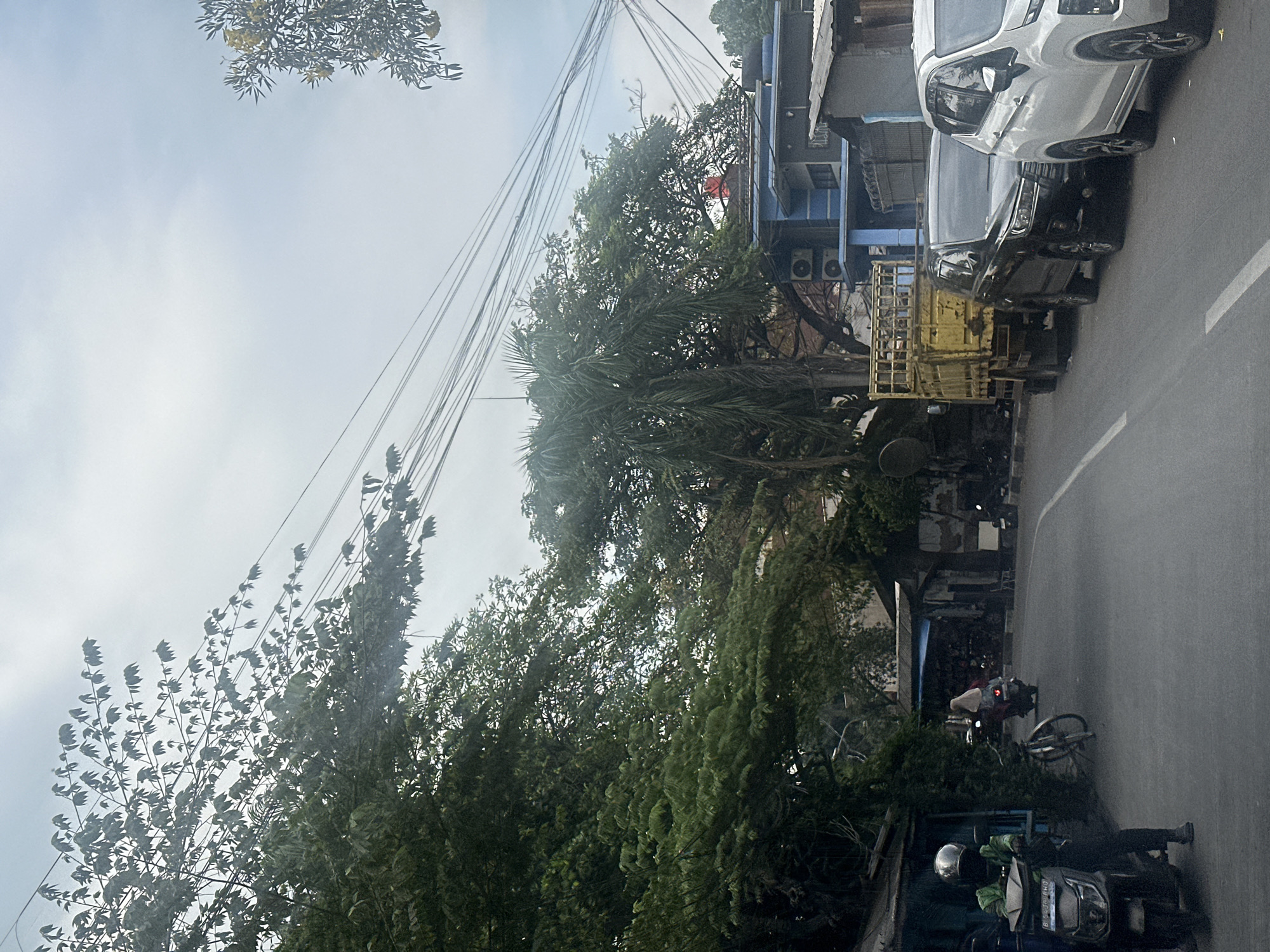 The image depicts a street scene in North Jakarta, Indonesia. A white car and a black SUV are parked next to a yellow truck along the side of the road. Power lines run above the street, obscured by trees. Two motorcycles are also visible on the road, each with riders. Buildings are seen on the right side of the frame.
