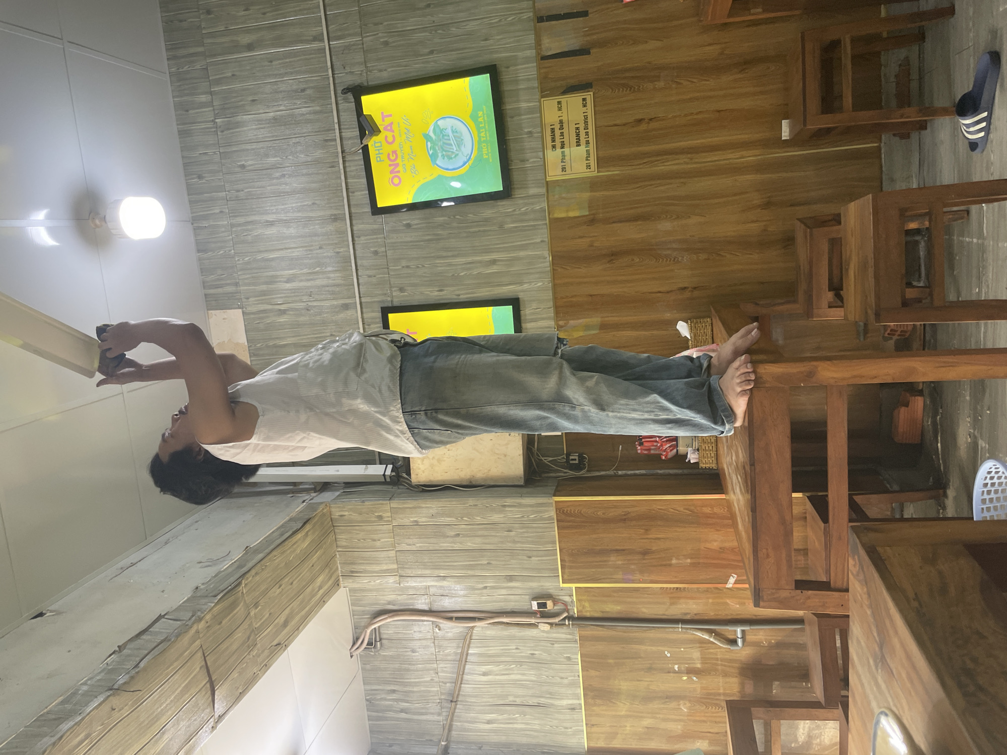 A man stands on a table in what appears to be a restaurant in Thủ Đức, Vietnam, reaching up to adjust a light fixture or fan on the ceiling. He is wearing a white sleeveless shirt and blue jeans. The restaurant has wooden paneled walls and furniture. There are also signs on the wall, one of which appears to be advertising pho. Several small wooden stools are visible around the tables. The overall environment suggests a casual dining establishment.