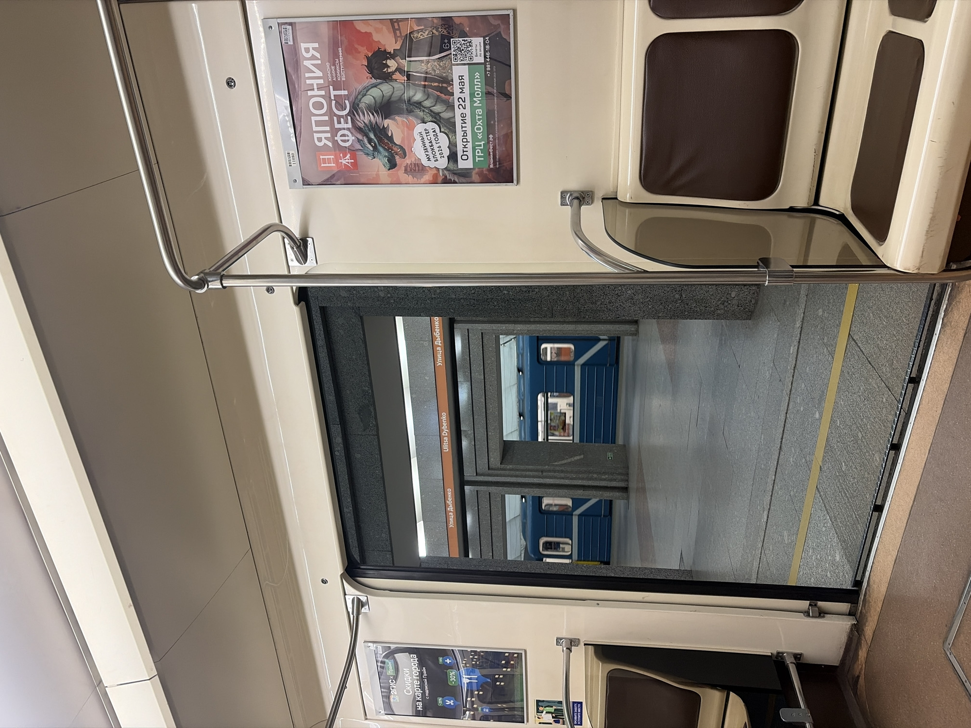 The image depicts the interior of a subway car in Saint Petersburg, Russia, as it arrives at the Ulitsa Dybenko station. The camera is positioned inside the subway car, facing the open doorway. Through the doorway, we can see the station platform and another subway train waiting on the opposite track. Signs identifying the station as "Ulitsa Dybenko" in Russian are visible above the platform entrance. Inside the car, there are beige wall panels, brown padded seats, and handrails. Two advertisements are visible on the wall of the subway car. One is for "Japan Fest," featuring a dragon and anime characters. Another is for discounts with 2GIS on the city map.