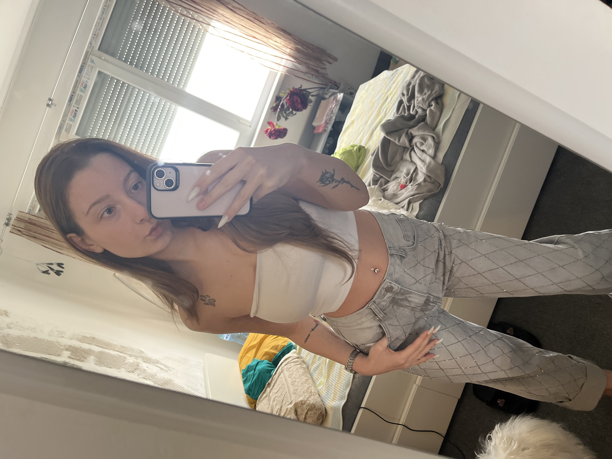 A young woman is taking a selfie in a mirror. She has long brown hair and is wearing a white tube top and light grey jeans with a diamond pattern. A navel piercing is visible. She is holding a white iPhone with multiple lenses in her right hand. Her left hand is resting on her hip. Tattoos are visible on her arm and shoulder.
Behind her in the reflection of the mirror, there's a bedroom. A bed with striped bedding is visible. There are flowers on a stand near the window. The window has blinds. On the floor, near the bed, there's a white fluffy dog.
No landmarks that indicate a location in Varna, Bulgaria are seen. The image appears to be taken indoors.