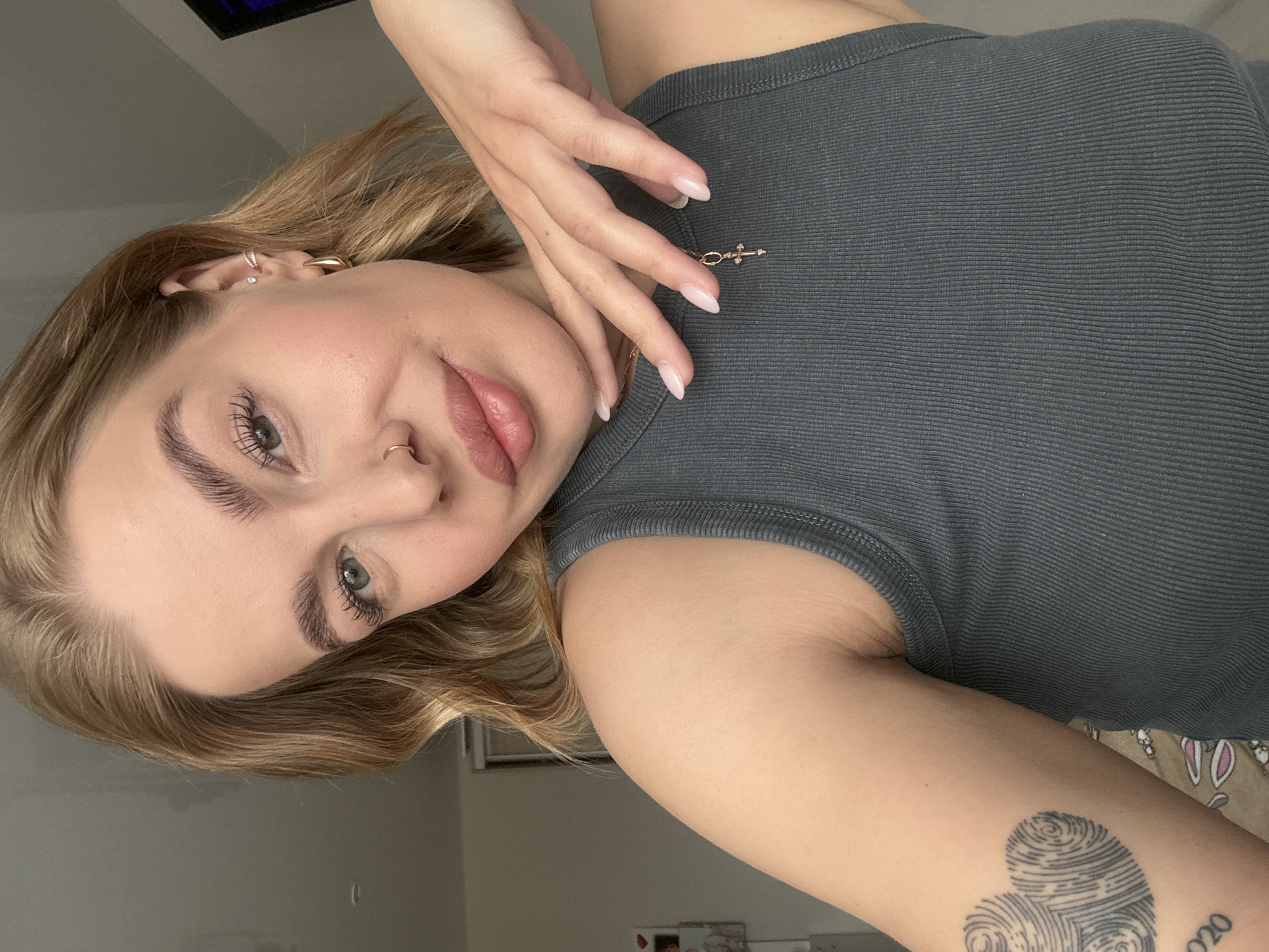 A young woman poses for a selfie. She is wearing a grey tank top and has a tattoo of fingerprint hearts on her right forearm, along with the text "2020". She has light blonde hair, light skin, and green eyes. She is wearing a nose ring, earrings, and a delicate cross necklace. Her nails are manicured with a light pink color. The background appears to be a room interior, with a glimpse of a television and some blurred items on a shelf. There is no visible indication of the location being Chaikovsky, Russia.