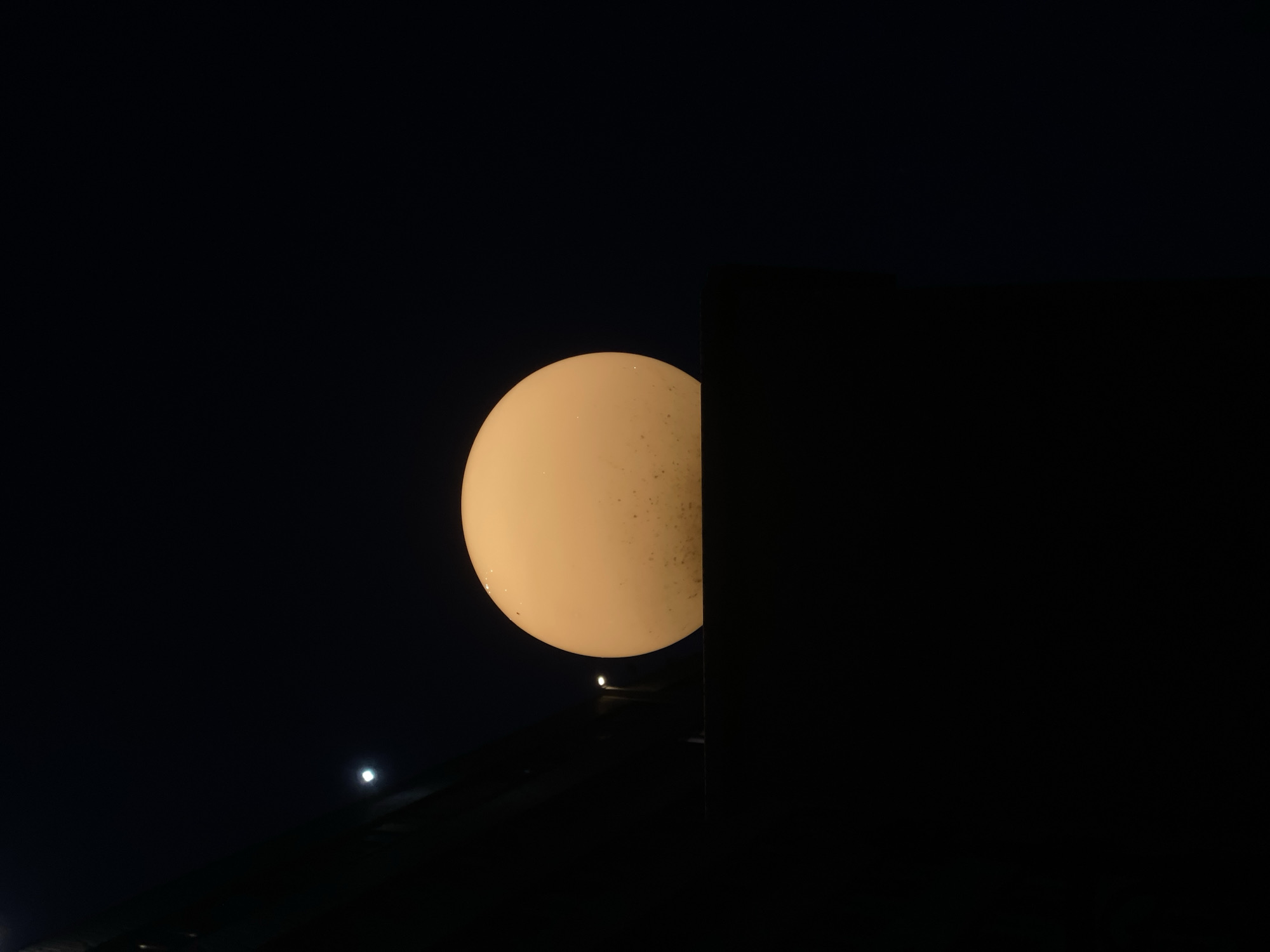 A low-angle, nighttime view shows a large, illuminated globe-shaped lamp partially obscured by a dark, rectangular structure on the right. A smaller light source is visible below and to the left of the globe, near a dark surface. A fainter light is further to the left in the dark background. The setting appears to be Chaikovsky, Russia.