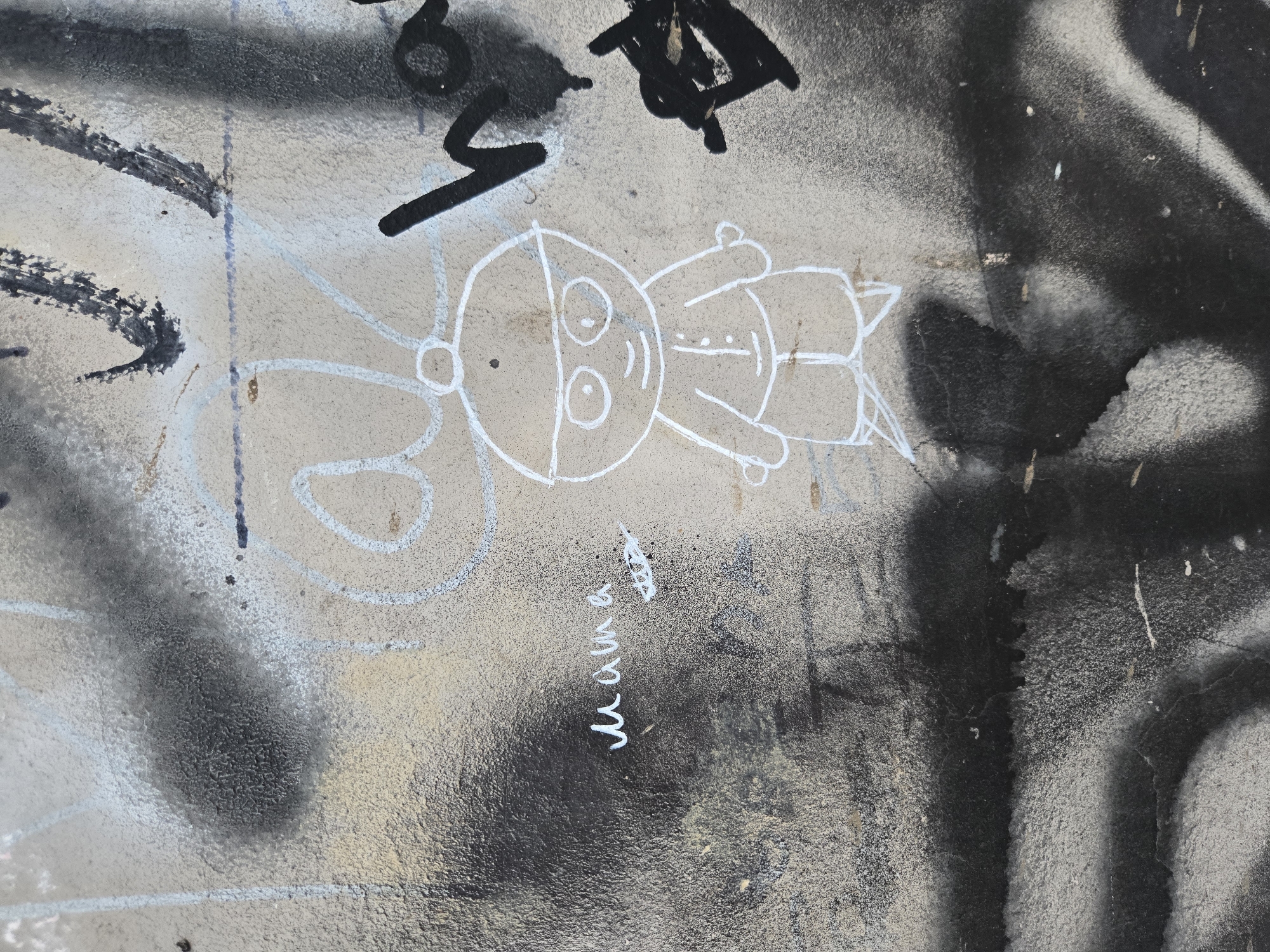This close-up image captures a segment of a textured wall, likely in Budva, Montenegro, adorned with various layers of graffiti. The focal point is a whimsical, childlike figure drawn in white outline, resembling a cartoon character or a doll. It features a large, round head with wide eyes and a small mouth, a simple body with outstretched arms, and legs. An antenna-like shape protrudes from the left side of its head, and a vertical line divides its face.
Surrounding and partially overlapping this white drawing are bold, black spray-painted strokes and abstract shapes, some indistinctly resembling letters or numbers, indicating earlier or more chaotic graffiti. Additional white lines and circular patterns are visible at the periphery, suggesting other incomplete drawings. The wall itself exhibits a mottled texture, with areas of light beige-grey and darker black-grey paint, interspersed with white paint splatters and drips, reflecting multiple layers of artistic expression and wear.
A notable detail is the handwritten word "mama" in white cursive, located below the main figure, accompanied by a small arrow pointing to the right. The image provides no specific cues for time of day or weather, but the combination of worn surfaces and layered street art creates an authentic urban aesthetic. The scene is a static display of public art, contrasting a simple, innocent drawing with more aggressive, abstract markings.