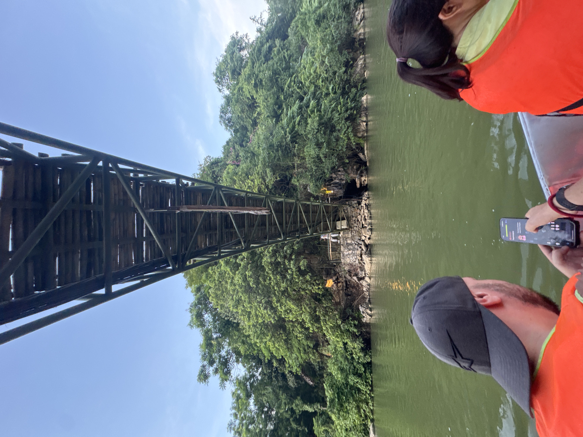 This is a photograph taken from a boat on a body of water, looking up at a large, wooden and metal bridge. The bridge appears to be an elevated walkway or pedestrian bridge that spans over the water and a vegetated cliff face. On the cliff face, there are several torches burning, casting an orange glow. A person in light-colored clothing is visible standing on a walkway near the torches. The water is green, and the surrounding environment is lush with dense green trees and foliage. In the foreground, two people are visible from behind, also on the boat. One person is wearing a black baseball cap and an orange life vest, and is holding a smartphone. The other person is wearing an orange life vest and has their hair in a ponytail. The sky above is a clear blue. The scene is likely located near the city of Yên Sơn Ward, Vietnam, given the architectural style of the bridge and the surrounding natural environment.