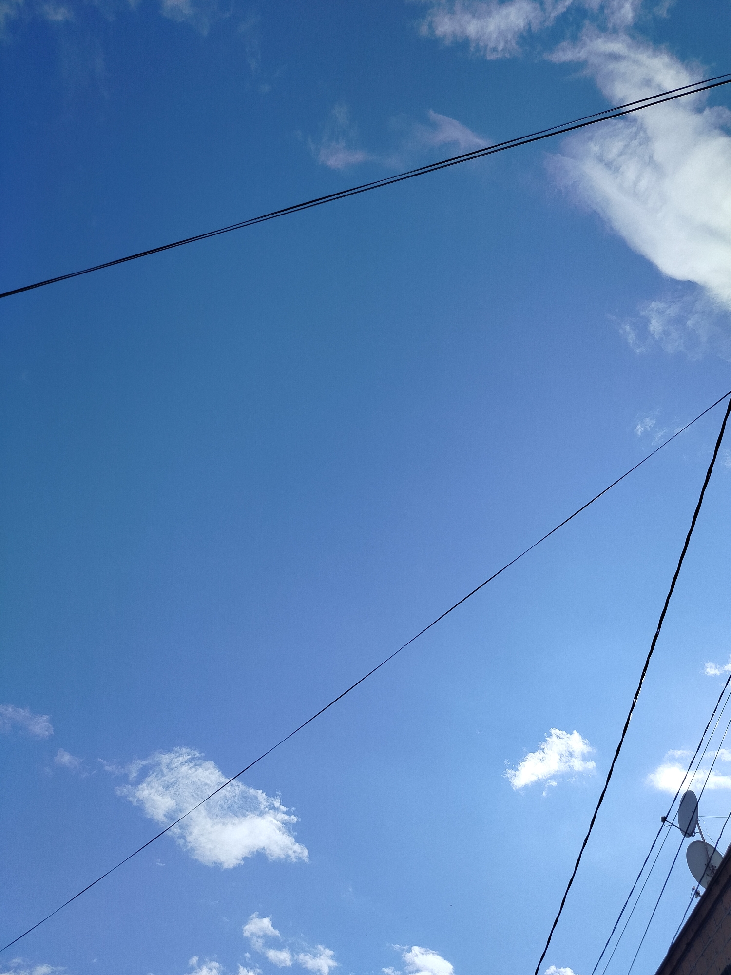 The image shows a section of the sky in Yerevan, Armenia, with a few clouds scattered throughout. Several electrical wires are visible, stretching across the frame. In the bottom right corner, a building with two satellite dishes mounted on its side is partially visible.