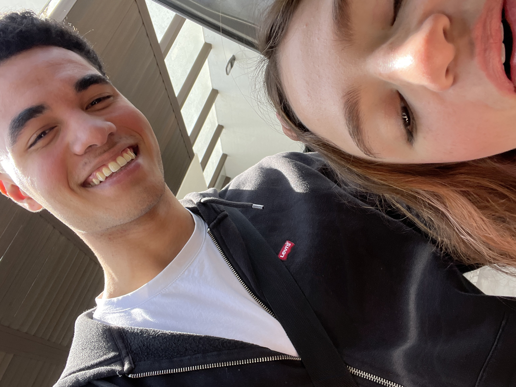 A selfie-style photograph captures a man and a woman, both appearing young, posing together outdoors. The man, positioned on the left side of the frame, is smiling broadly, revealing his teeth. He has dark hair and is wearing a white t-shirt under a black, zippered hoodie. The hoodie features a visible "Levi's" tag on the chest. The woman, tilted at an angle on the right side, also appears to be smiling with her mouth open. She has light brown hair. The background suggests an urban environment with building structures visible. The photograph was taken at an angle, looking upward, capturing the subjects against the backdrop of the environment. There is no indication this photograph was taken in San Bernardino Tlaxcalancingo, Mexico.