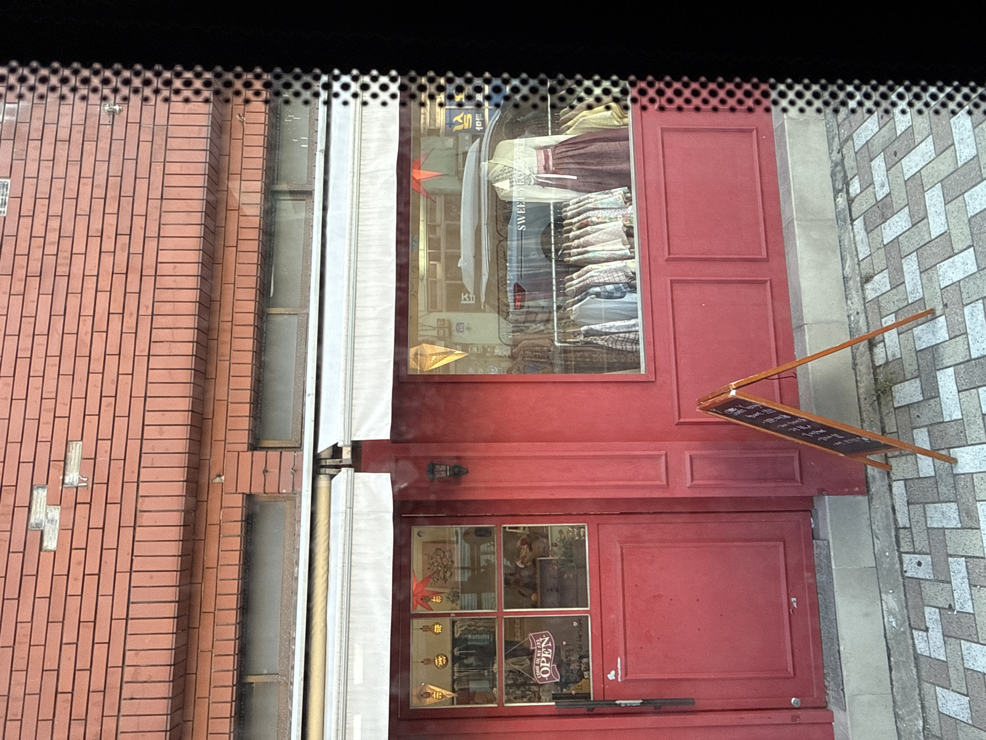 The image depicts the exterior of a red-fronted shop with large windows and a red door. Above the windows, a red brick wall is visible, showing some signs of wear. The shop windows display clothing, including what appears to be a hanbok (traditional Korean garment) on a mannequin. A rack of shirts is also visible in the window. A small sign on the door reads "COME IN WE ARE OPEN". An A-frame chalkboard sign stands on the sidewalk in front of the shop. The ground is paved with a brick pattern. The scene appears to be a street in an urban environment.