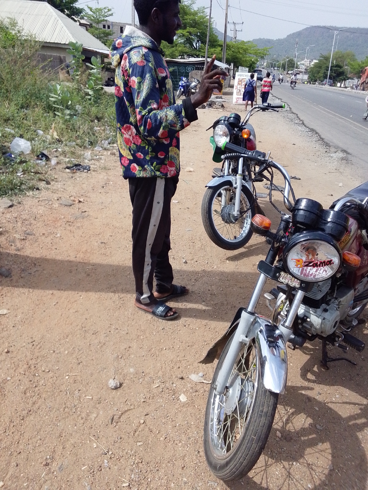 The image depicts a man standing next to two motorcycles on a dirt road in Adankolo, Nigeria. The man is dark-skinned, wearing a floral-patterned jacket, dark pants with a stripe down the side, and sandals. He appears to be holding a small object in his hand. Two motorcycles are parked near him on the dirt road; the closer one has the name "Zama" on the headlight. In the background, there is a paved road with some people walking, a building with a corrugated metal roof, and some trees and a hill in the distance. Trash is scattered on the ground near the man.