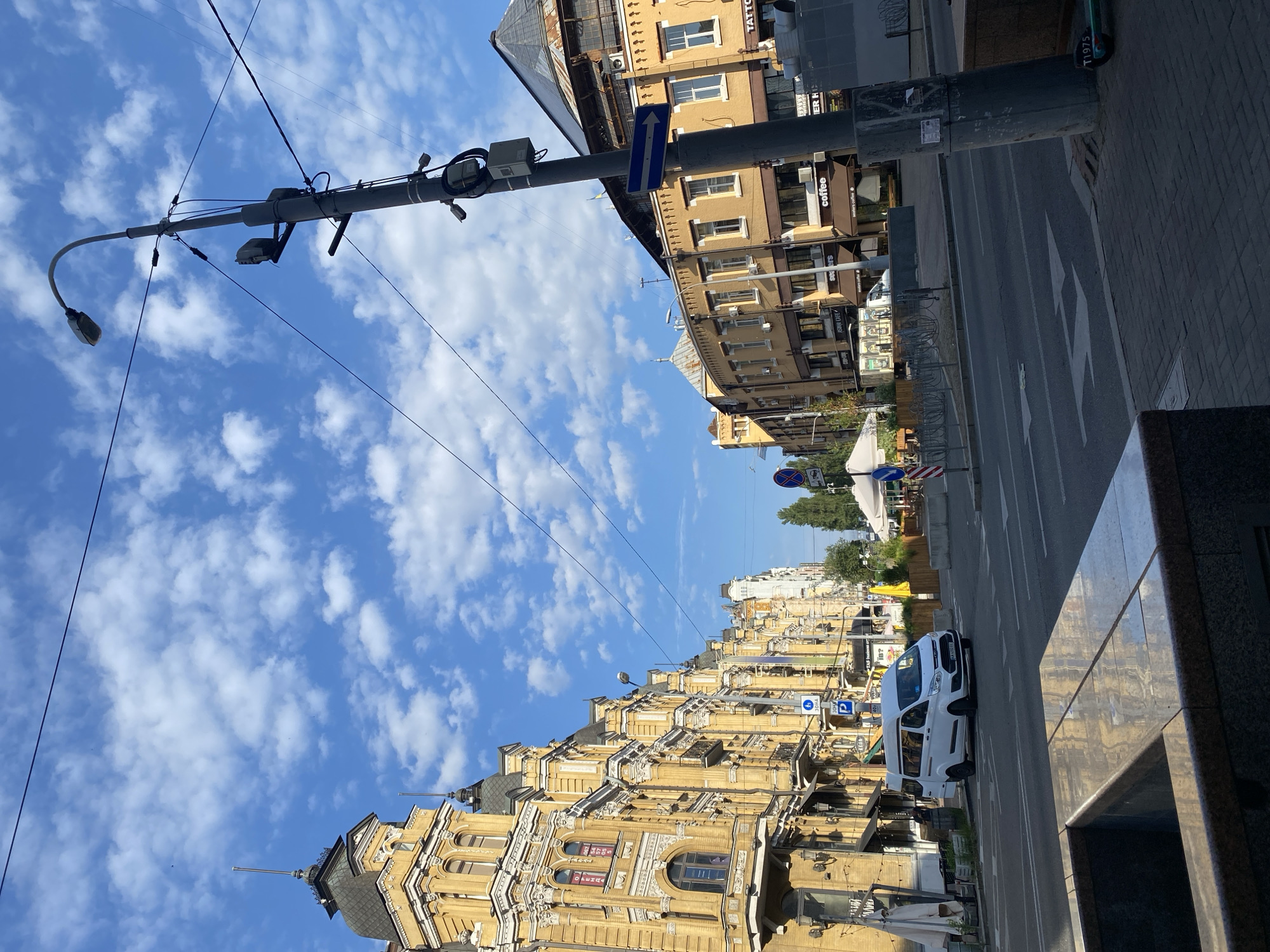 The image captures a street scene in a city, likely during daytime given the bright sky and shadows. The architecture on the left is ornate and yellow, with a detailed facade and a pointed roof. Across the street, buildings are also yellow and brown, with storefronts visible, including signs for "coffee," "asian cafe," and "TATTO." A white van is driving on the road, and several traffic signs are present, including directional arrows and a parking symbol. A lamppost with attached electrical equipment stands prominently in the foreground on the right. The sky is blue with scattered white clouds. The street is paved, and a sidewalk with a stone border is visible. The overall impression is of a moderately busy urban environment with older, European-style buildings.
