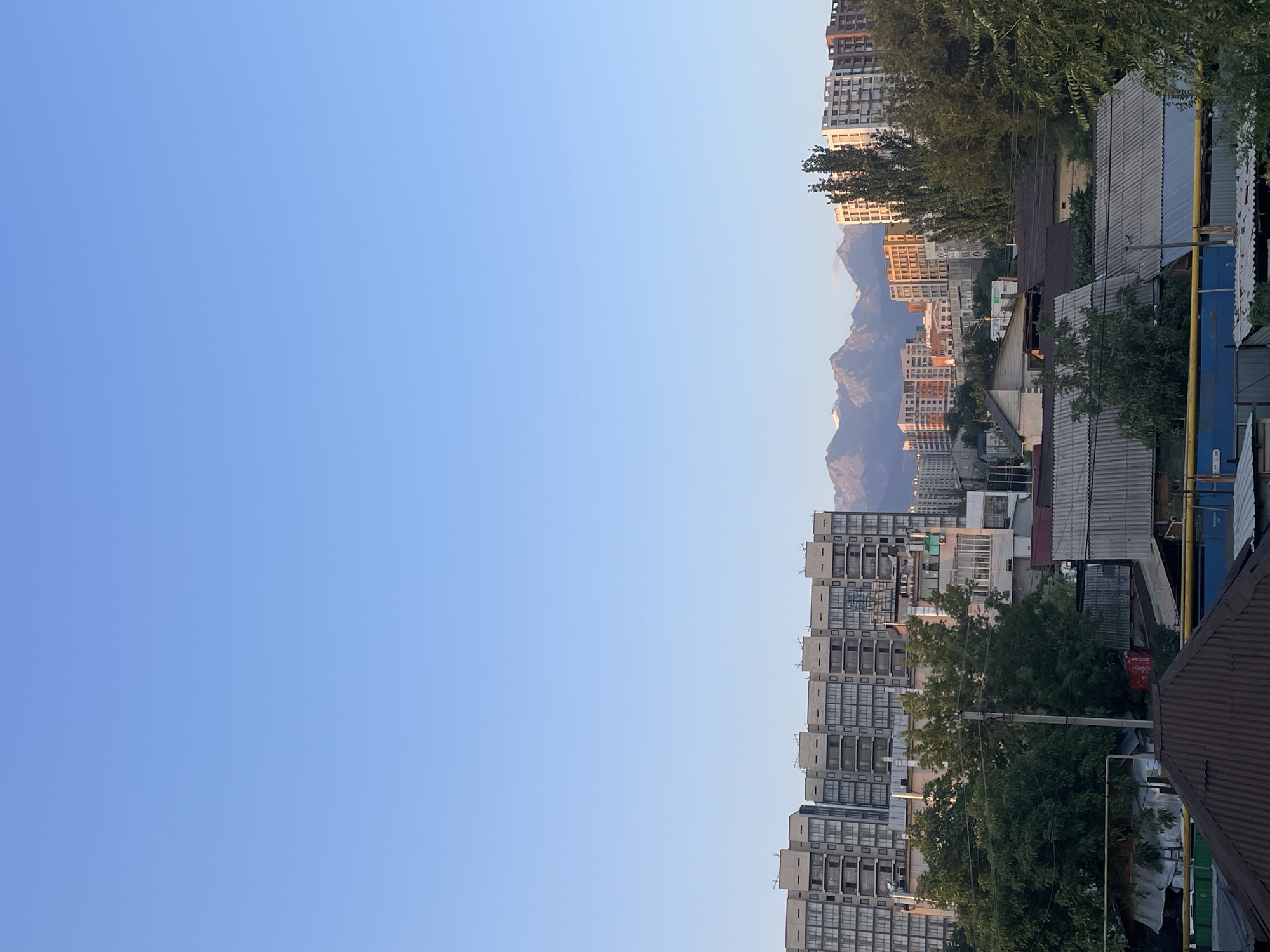 The image depicts a panoramic view of a city skyline, with a prominent mountain range in the background. The foreground features a collection of low-rise buildings, including houses with corrugated metal roofs, suggesting a more residential or older part of the city. Above these, several mid-rise and high-rise apartment buildings are visible, some appearing modern with balconies and glass facades, while others seem to be under construction or are older in design. The distant mountains are snow-capped, indicating a significant elevation and a clear, bright atmosphere. The sky is a clear, pale blue, transitioning to a slightly warmer tone near the horizon, suggesting it might be late afternoon or early morning. Trees are scattered throughout the scene, particularly in the mid-ground, providing a contrast to the urban structures. The overall impression is that of a city nestled at the foot of a majestic mountain range, a common characteristic of Almaty, Kazakhstan.
