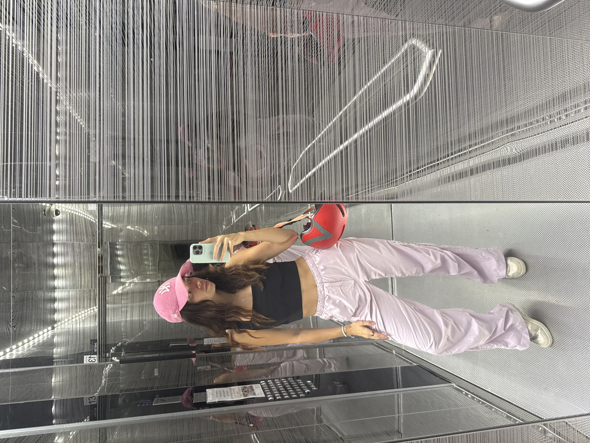 A young woman poses for a selfie in what appears to be an elevator or lift, as indicated by the mirrored interior and metal paneling. She is wearing a black crop top, lavender parachute pants, a pink baseball cap, and white sneakers. She holds a smartphone in her right hand to take the picture and has a red helmet resting on her arm. The background features a modern aesthetic with vertical metal stripes and reflective surfaces, creating multiple mirror-image effects. Text such as "13" is partially visible on the side of the elevator wall. The location in Tel Aviv, Israel is not discernible from the image.