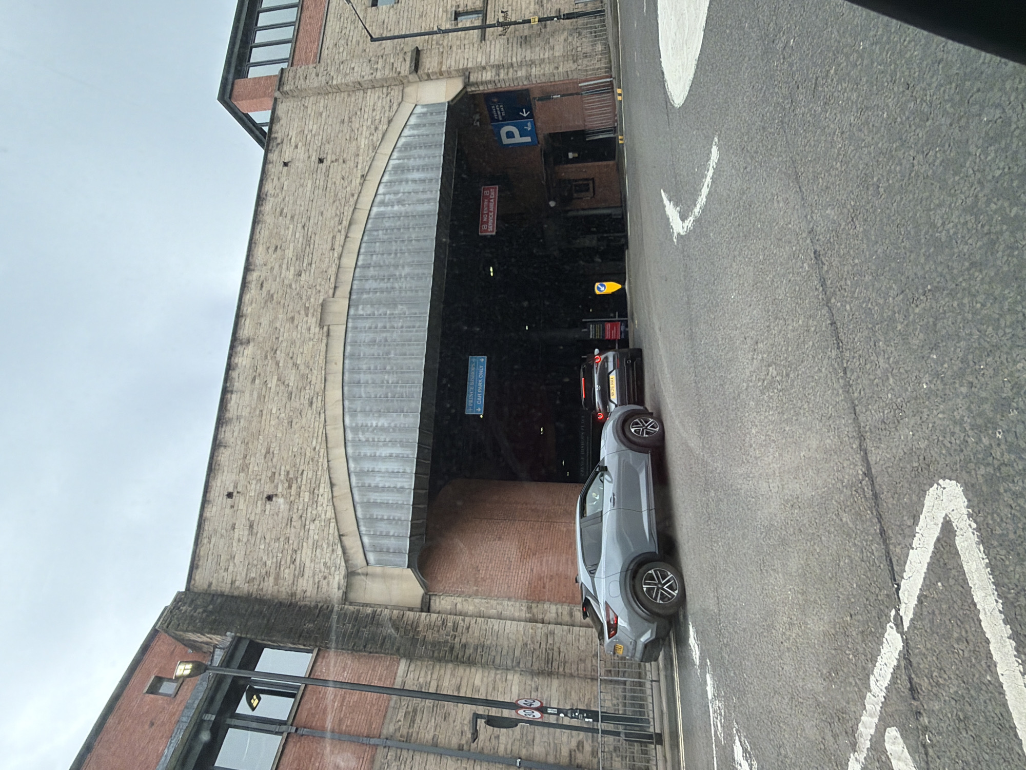 The image shows a street scene in Durham, United Kingdom. A light gray Nissan Juke is parked on the right side of the road, adjacent to a large stone archway that serves as the entrance to the Prince Bishops Place car park. The road is marked with white painted lines, including chevrons and directional arrows. Above the car park entrance, a blue sign with a "P" indicates parking. Red and white circular speed limit signs are mounted on a pole in the lower left foreground. The architecture suggests an older building, likely sandstone, with modern windows on the upper levels. The sky is overcast. Another vehicle, black, is partially visible in front of the Juke in the parking entrance.