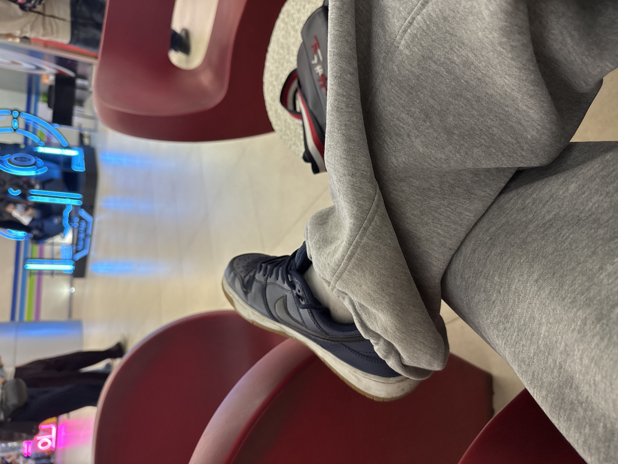 The image, taken in Yerevan, Armenia, shows a close-up, rotated view of a person's lower body in an indoor setting. The main subject is a leg clad in grey sweatpants or joggers, with a dark blue Nike Dunk sneaker on the foot. A white sock is visible between the pant cuff and the shoe. The foot is resting on a curved, red piece of furniture, possibly a bench or chair, which dominates the foreground.
Behind the leg, a grey backpack with red text, appearing to be Japanese script "カナダ" (Kanada), is partially visible. The background is a brightly lit, blurred indoor space. To the left, vibrant blue neon lights form abstract patterns or a user interface on what might be an arcade machine or a decorative display. Further in the background, out-of-focus figures of other people can be seen, suggesting a public environment. A blurry pink neon sign is also visible in the distant background, displaying indiscernible symbols that resemble East Asian script.
The scene depicts a casual moment in a modern commercial or entertainment complex, such as a mall or arcade, characterized by contemporary furniture and neon lighting. No specific activity beyond sitting is clearly identifiable. The time of day is not evident due to the artificial lighting.