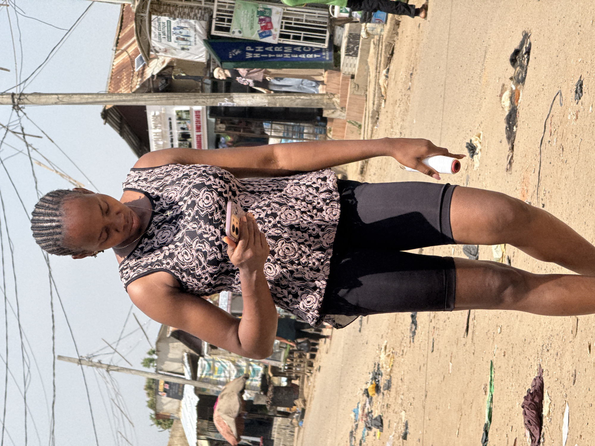 A young Black woman with braided hair is holding a spool of white thread in her right hand and a mobile phone in her left. She is wearing a black and pink floral patterned top and black biker shorts. She appears to be looking down at her phone. The scene is outdoors on a dirt road. In the background, there are buildings, a pharmacy sign, power lines, and scattered debris on the ground. The time of day appears to be daytime, and the weather seems clear. The overall setting suggests a busy street or marketplace in Abuja, Nigeria. Visible text includes "LUXURY COSMETIC & BOUTIQUE" and "PHARMACY".