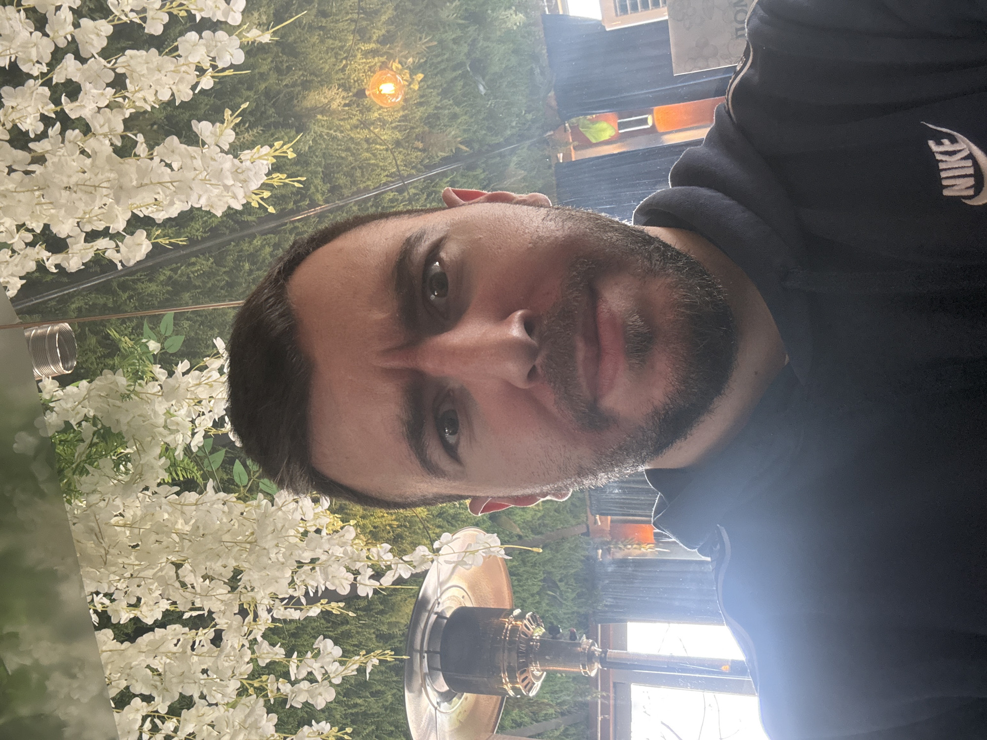 A close-up portrait shows a man with dark hair and a beard wearing a dark-colored Nike hooded sweatshirt. Behind him is a floral arrangement consisting of white flowers. A patio heater is visible in the lower center. It's unclear whether this image was taken in Sofia, Bulgaria.