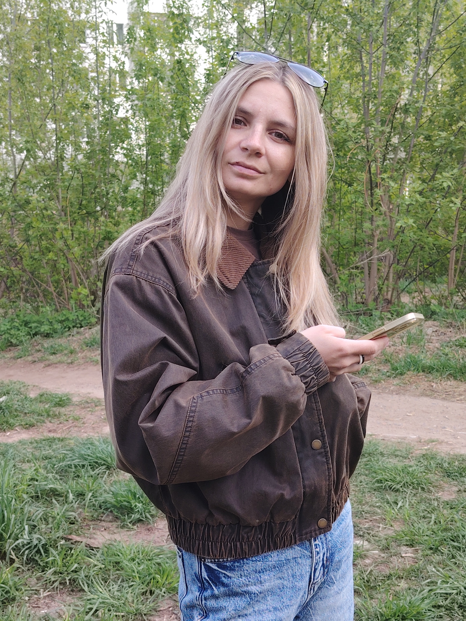 A woman is standing outdoors, holding a gold-colored phone. She has shoulder-length blonde hair and is wearing blue jeans, and a brown jacket with a corduroy collar. She has a pair of glasses resting on top of her head. The background includes trees and a dirt path. The location is Chaikovsky, Russia.