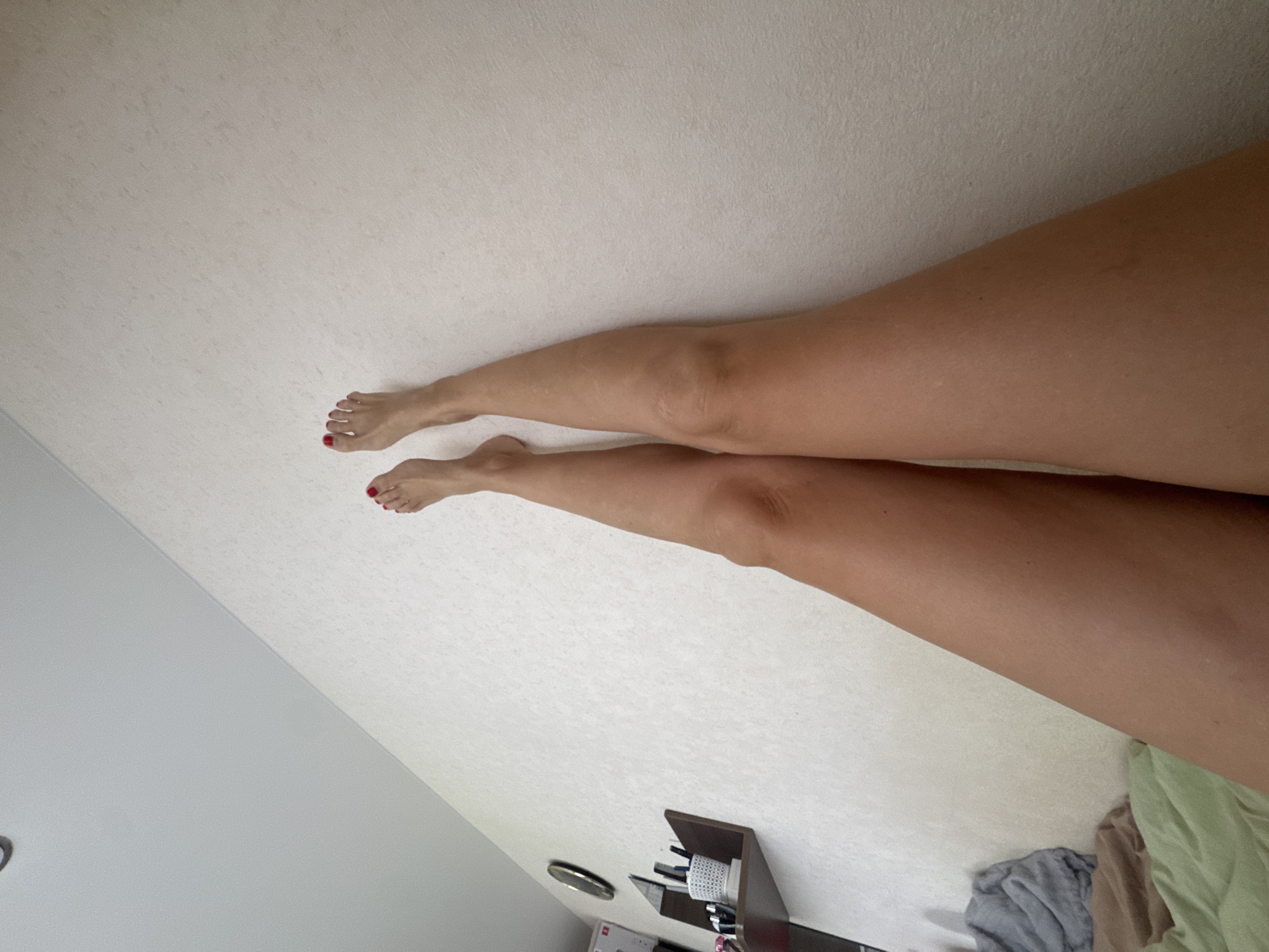 The image shows a view of a person's legs from a slightly downward-looking perspective. The person is lying on a bed with white patterned bedding, and the legs are stretched out towards the top left of the frame. The feet are visible, with red nail polish on the toes. A small shelf with various items is visible in the lower part of the image.
The location of the image is unknown. There is no information to indicate that the location of the image is in the city of Vesyoloye, Russia.