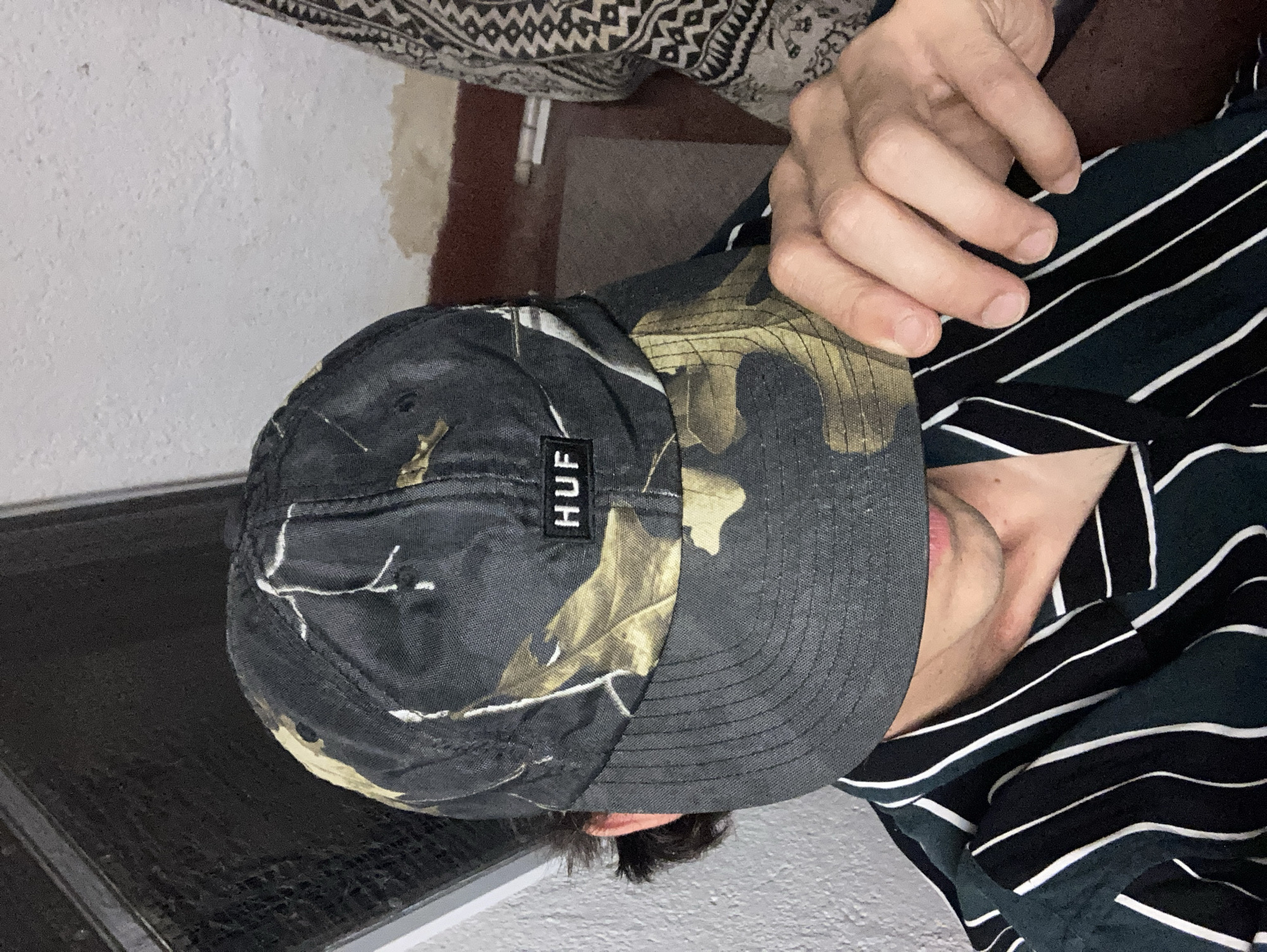 A person is seen from the chest up, wearing a camouflage patterned baseball cap that partially covers their face. A label with the letters "HUF" is visible on the cap. The person is also wearing a dark green and white striped shirt. Their hand is raised, resting on the brim of the cap. The background appears to be an indoor setting with textured walls and a dark window or frame on the left. There is a patterned fabric visible on the right side of the image, suggesting another person or furniture.