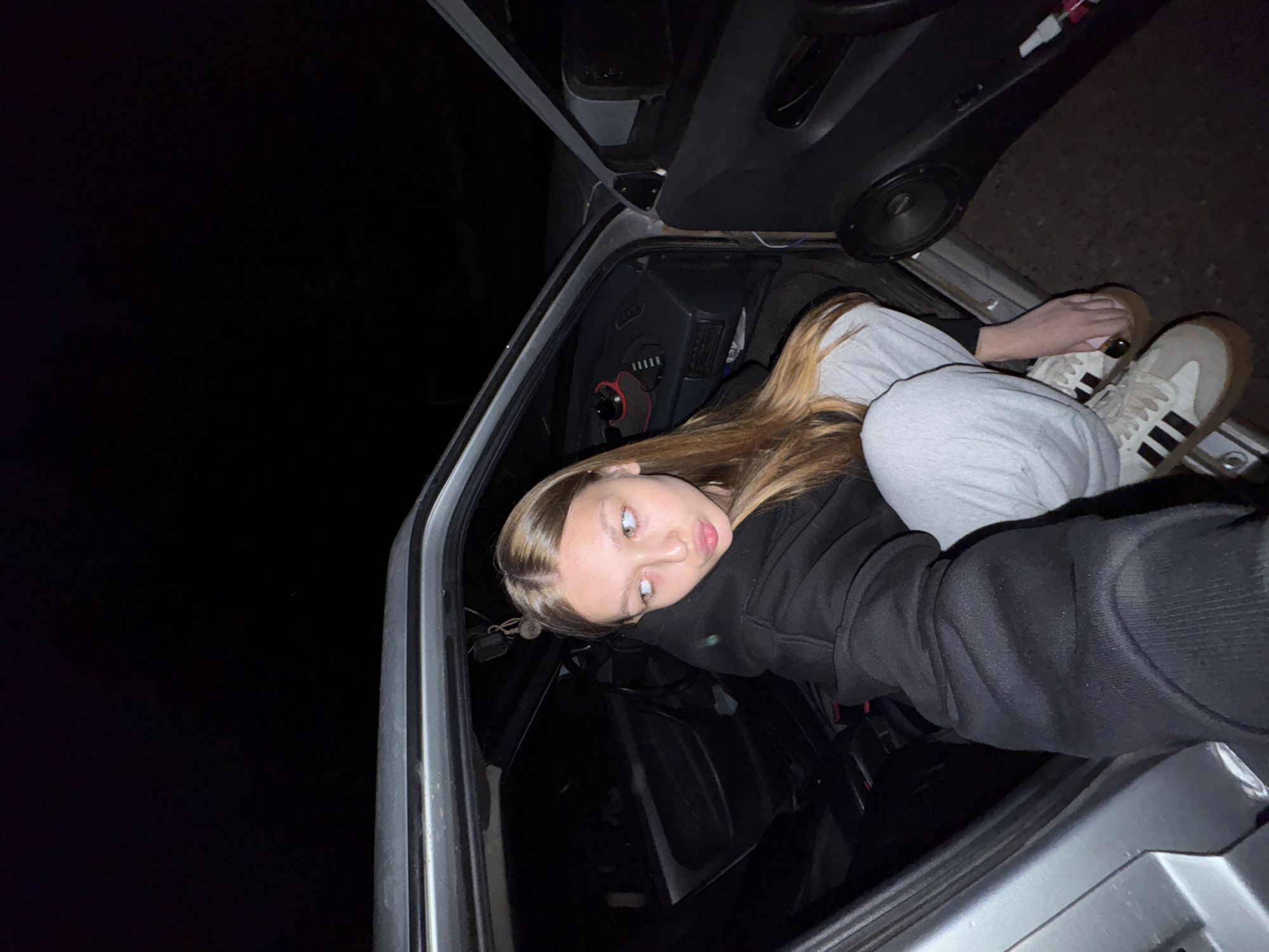 An image captured at night in Chaikovsky, Russia, features a young adult female partially inside the front passenger side of a silver or grey sedan. The woman, with long, straight, light brown hair, has light-colored eyes and her lips are pursed in a "duck face" expression as she looks to her right (viewer's left). She is wearing a black hooded sweatshirt, light grey sweatpants, and beige/white Adidas Samba sneakers with brown gum soles and black stripes. Her left arm (viewer's right) is extended, holding a device to capture the image, consistent with a selfie. Her right leg is positioned outside the car, while her left leg is inside, suggesting she is either entering, exiting, or posing.
The car's interior is visible, featuring a dark-colored dashboard with gauges and a red and black device attached. A speaker is integrated into the lower panel of the front passenger door. Both the front and rear passenger doors are open. A small dangling ornament is visible near the rearview mirror area. A white spray bottle with pink accents rests on the ground near the open front passenger door. The background outside the vehicle is completely dark, indicating a nighttime setting with minimal external lighting. The ground directly beneath and around the car appears to be a dark, unpaved surface or asphalt.