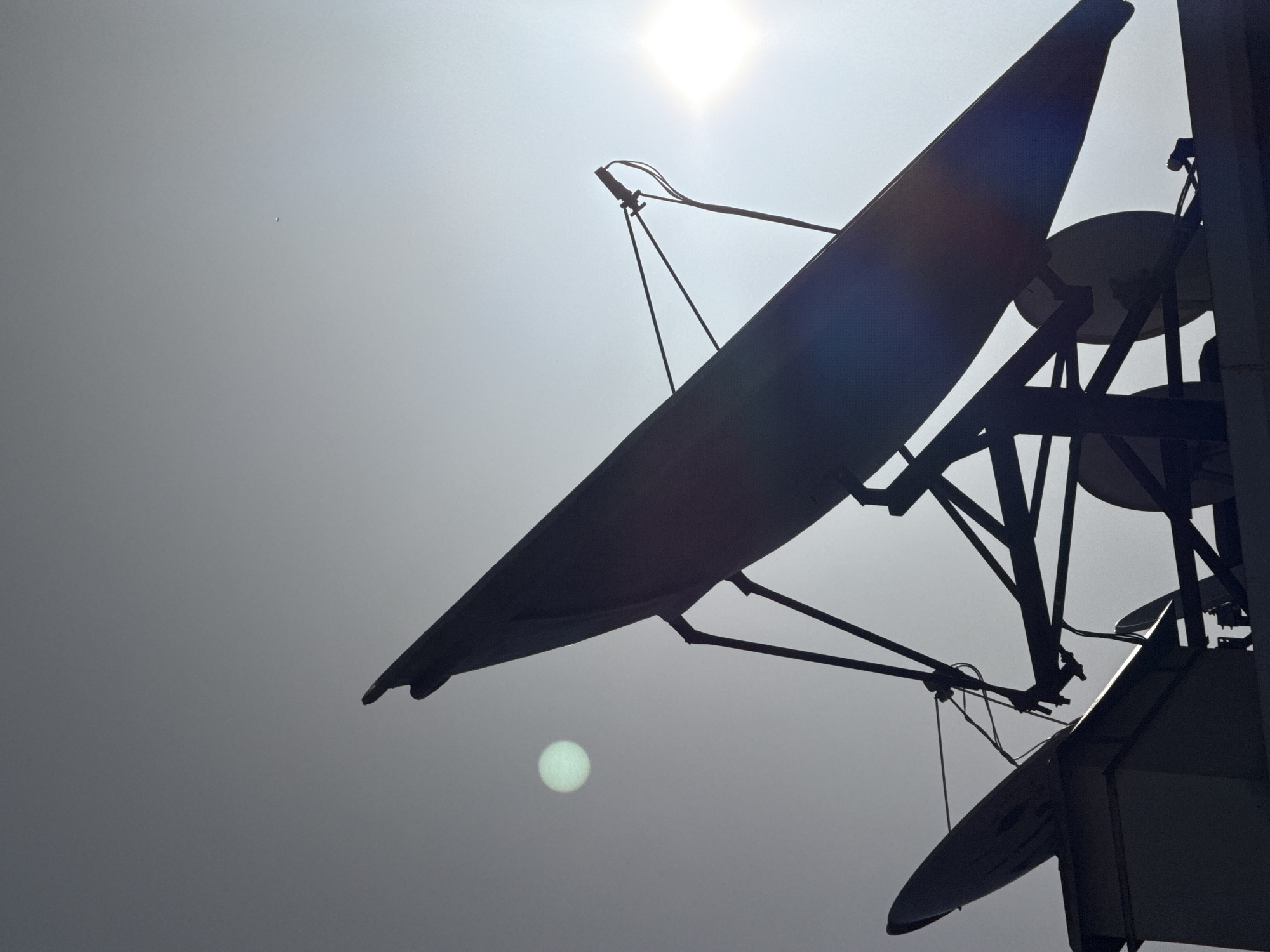 The image depicts a low-angle view of a satellite dish array against a bright, overcast sky with the sun prominently positioned in the upper center. The dishes are silhouetted, revealing their structure and support beams. A small, round halo effect is visible lower in the frame. The photograph does not include any people, specific subjects or actions beyond the inherent presence of the satellite equipment. The precise location within Alanya, Turkey cannot be definitively determined from the image.