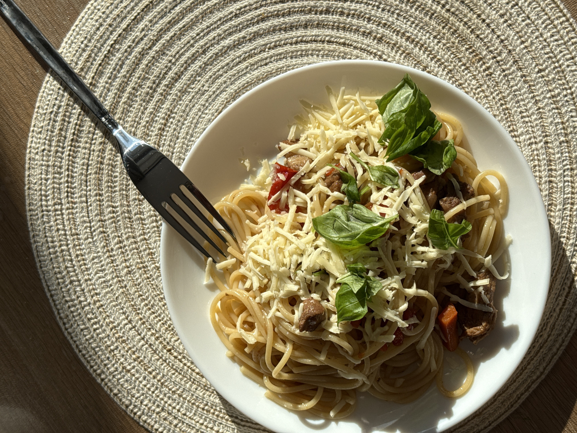 The image displays an overhead, close-up view of a serving of spaghetti on a white, round plate. The plate is positioned on a light-colored, circular placemat with a distinct woven texture. Strong, direct sunlight illuminates the scene, casting sharp shadows on the plate and placemat, indicating it is daytime with clear weather.
The spaghetti noodles are generously topped with shredded cheese and several vibrant green basil leaves. Mixed within the pasta are visible chunks of what appears to be cooked meat, possibly beef, along with small pieces of red vegetables, likely diced tomatoes or bell peppers.
A dark-colored metallic fork is placed on the left side, resting partially on the placemat and partially submerged in the spaghetti, suggesting the meal is either about to be eaten or is already being consumed. A segment of a wooden table surface is visible at the bottom left, peeking from under the placemat. There are no people present, and no visible text. The overall impression is a simple, appetizing homemade meal, located in Perm, Russia.