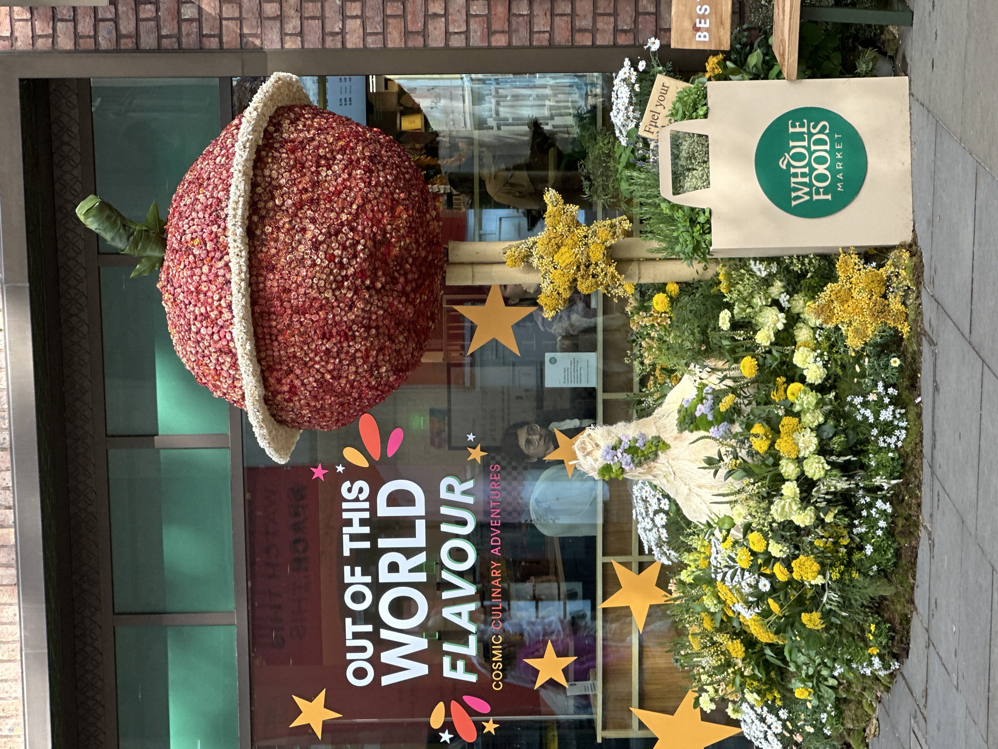 The image shows the exterior of a Whole Foods Market store, located in the city of London, United Kingdom. The storefront is decorated with a large, spherical ornament covered in red and white flowers, resembling a stylized fruit. The store window features a graphic that reads "OUT OF THIS WORLD FLAVOUR COSMIC CULINARY ADVENTURES," adorned with star shapes and colorful accents. To the right of the floral fruit is a floral arrangement. There is a brown Whole Foods Market shopping bag in the window display. The storefront is set against a brick facade. There is a reflection of people in the window. The image is vertically oriented.