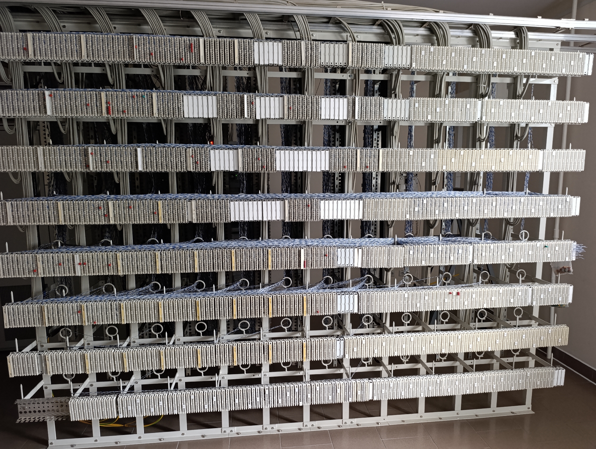 The image shows a large-scale telecommunications equipment rack in what appears to be a server room. The rack is filled with rows of modular connectors and wiring, organized in a highly structured manner. Thick gray cables run overhead, feeding into the rack. The equipment is predominantly white or light gray, with some visible wiring in blue, black, and red. There are no people visible in the image. The environment appears to be an indoor industrial or technical space, with a tiled floor and a plain wall visible on the right. The overall impression is one of complex and organized infrastructure for handling a significant volume of communication signals. The location is specified as the city council of Dubăsari, Moldova.