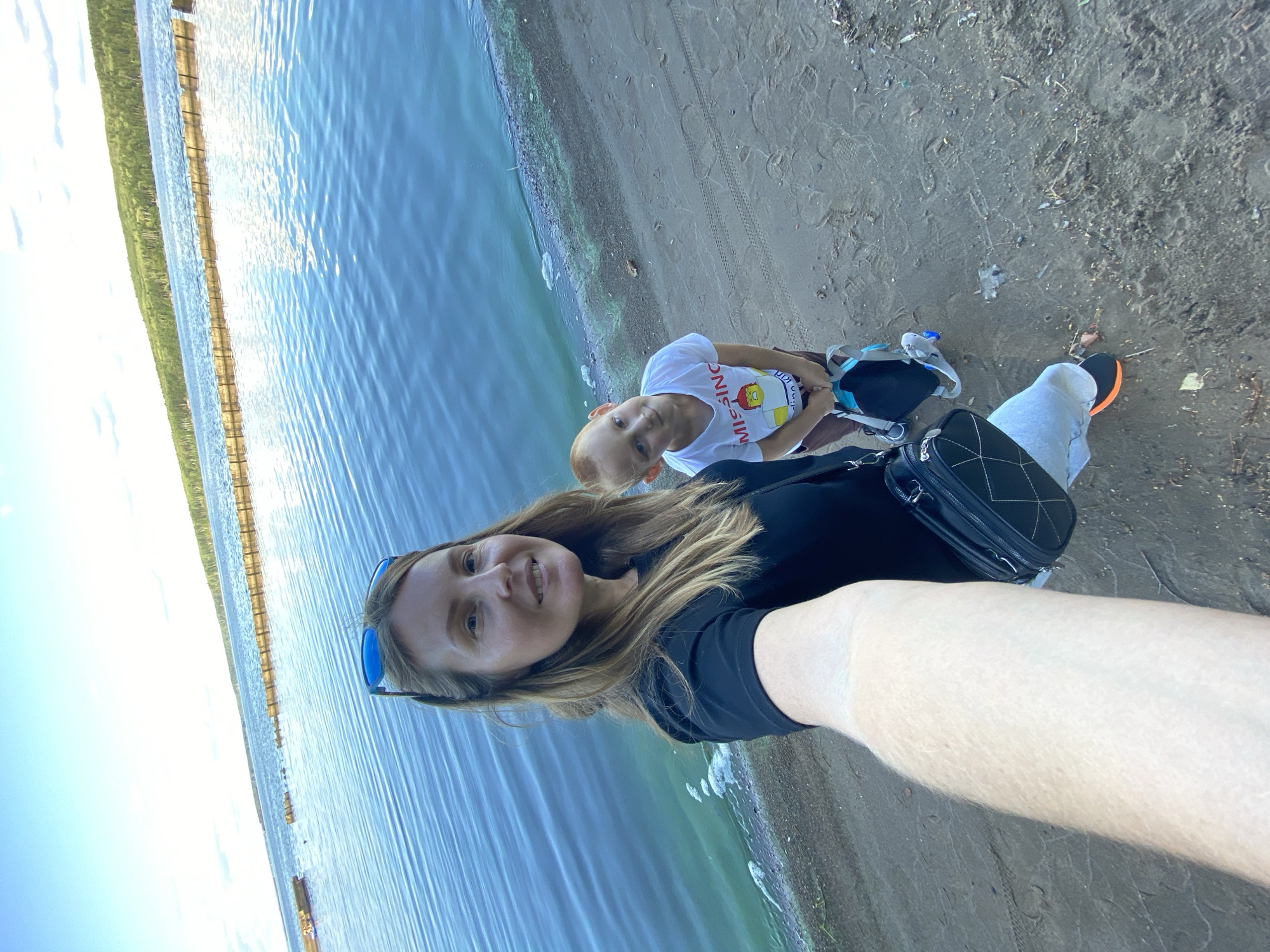 In an outdoor, high-angle, selfie-style shot, a woman with blonde hair and sunglasses on her head smiles at the camera. She is wearing a black t-shirt and a black crossbody bag. Beside her, to the right, a young boy with red hair and a white t-shirt that reads "MISSING" is holding a blue and black backpack. They are standing on a sandy shore next to a large body of blue water. In the background, a wooden structure extends into the water, and a tree-covered shoreline is visible under a bright, partly cloudy sky. The overall scene appears to be a natural setting, possibly a lake or a wide river.