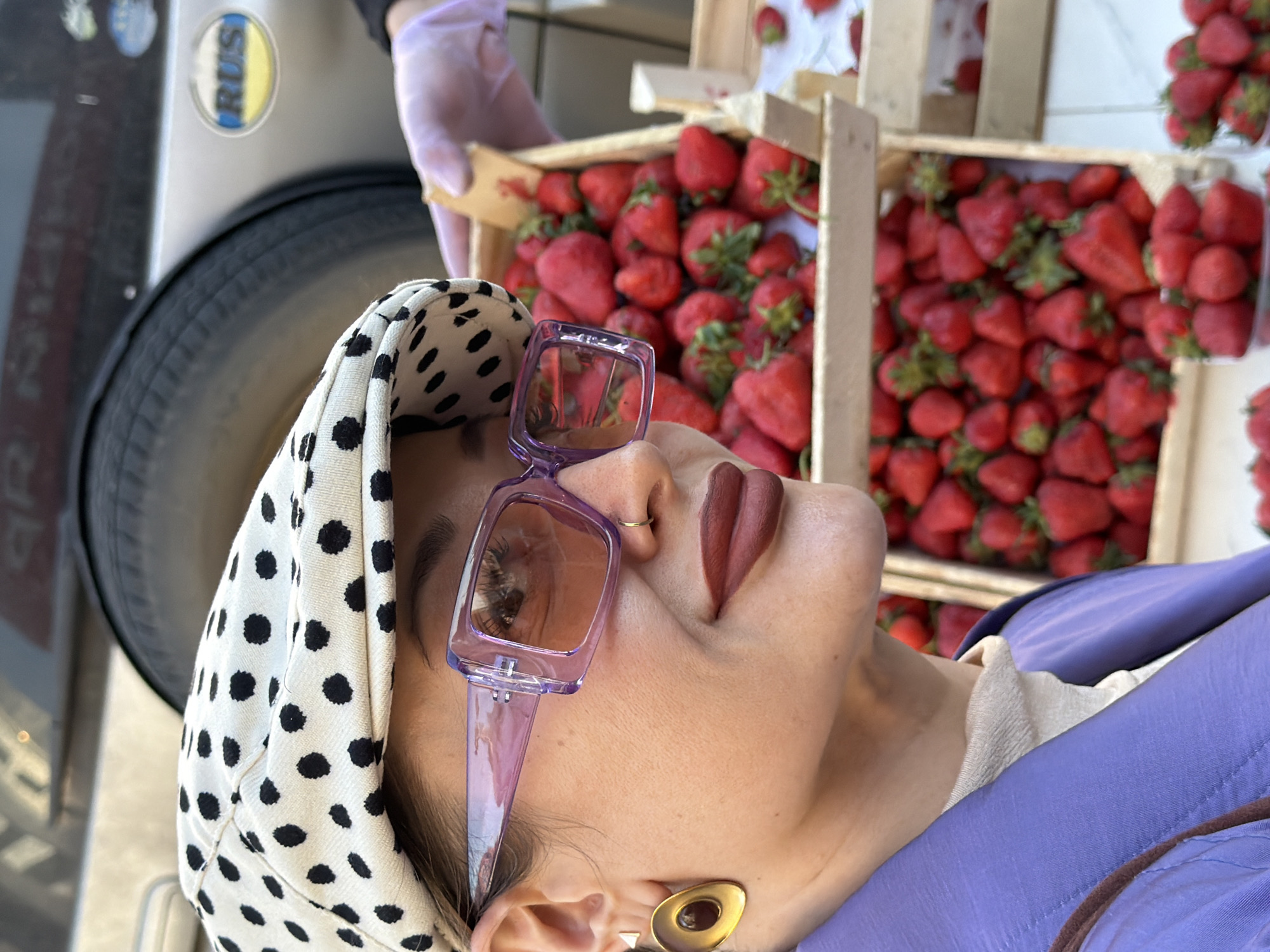 This image presents a close-up, angled view of a person surrounded by crates of fresh strawberries, set in an outdoor market context, likely in Krasnoyarsk, Russia.
The main subject is a person, likely female, whose head is tilted upwards and to the right, revealing their profile from the chin up. They wear distinctive, large transparent purple-framed sunglasses with brown tinted lenses. Their head is covered by a white headscarf or hood featuring black polka dots. The person is also wearing dark red matte lipstick and a small golden ring piercing on their right nostril. A prominent gold-colored, abstract-shaped earring adorns their right earlobe. They are dressed in a lavender or light purple jacket over a lighter-colored top.
The background is dominated by numerous wooden crates overflowing with bright red, ripe strawberries, creating a vivid and appealing display. A hand, encased in a light purple transparent glove, is visible in the upper part of the image, actively holding one of these wooden strawberry crates. This suggests an ongoing activity of selling, preparing, or selecting the fruit.
To the far left of the frame, a portion of a vehicle is visible, including its tire and side panel. A notable detail on the vehicle is a circular sticker displaying the text "RUS" in blue lettering on a yellow background, providing a clear location cue consistent with the provided context of Russia.
The lighting appears bright and natural, indicating that the scene takes place during the daytime. The overall atmosphere is vibrant and focused on fresh produce, characteristic of a market or street vendor setting. The person in the foreground could be a customer admiring the strawberries or a vendor at their stall.