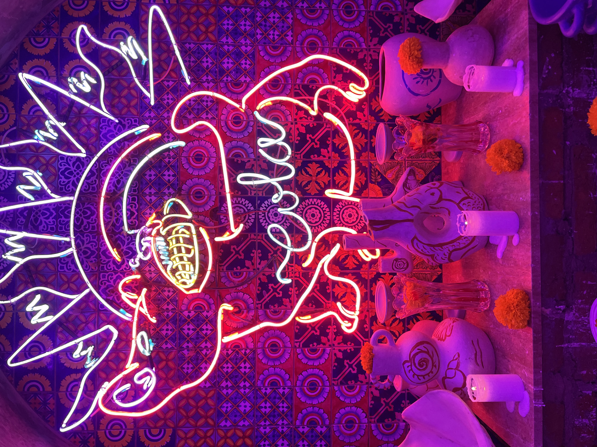 The image captures a decorated space in North Kuta, Indonesia. The background consists of a tile wall with intricate patterns of circles and crosses, predominantly in shades of blue, purple, red and orange. A neon sign depicting a bird or mythical creature is prominently displayed against the tile wall, outlined in bright orange and white neon lights.
To the right of the neon sign, there are several decorative items arranged on a surface. These include white ceramic skulls with painted designs, glass vases filled with orange flowers, and lit white candles. The light in this area is predominantly purple, creating a vibrant, yet slightly eerie, ambiance.