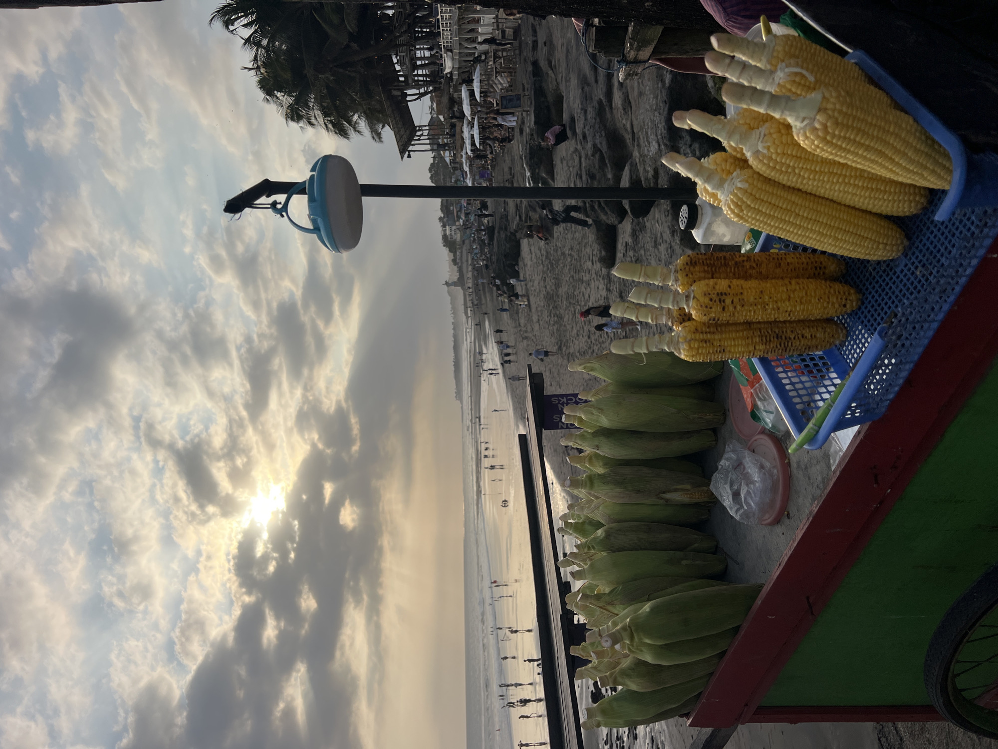 This image captures a vibrant scene on a beach in North Kuta, Indonesia, likely during sunset. In the foreground, a colorful cart laden with fresh corn is prominently displayed. Some of the corn is still in its husks, while others have been shucked and appear cooked, arranged in a blue basket. The sand-covered beach stretches into the distance, with numerous people scattered along the shoreline and wading in the ocean. The water reflects the soft glow of the setting sun, which breaks through a dramatic sky filled with clouds. Palm trees and some structures, possibly cafes or restaurants, are visible further down the beach, hinting at the lively atmosphere of the location. A street lamp with a distinctive blue light fixture stands tall on a pole, adding to the ambiance of the beachside setting.