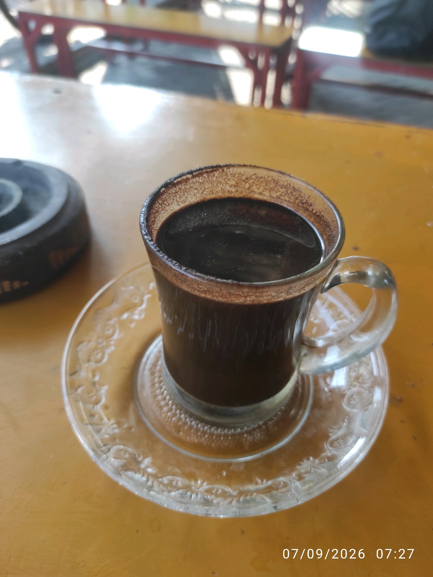 A close-up shot captures a clear glass mug filled with dark, hot coffee, resting on a clear glass saucer. The mug has a textured rim, likely from coffee grounds. To the left of the mug, a dark, round ashtray is visible. The objects sit on a worn, yellow wooden table. In the blurred background, a red and yellow striped table with chairs can be seen, suggesting an outdoor or semi-outdoor dining area. A timestamp in the bottom right corner reads "07/09/2026 07:27", indicating the time and date the photograph was taken. The scene is likely set in a casual eatery or coffee shop in Surabaya, Indonesia, given the presence of local-style coffee and the overall ambiance.