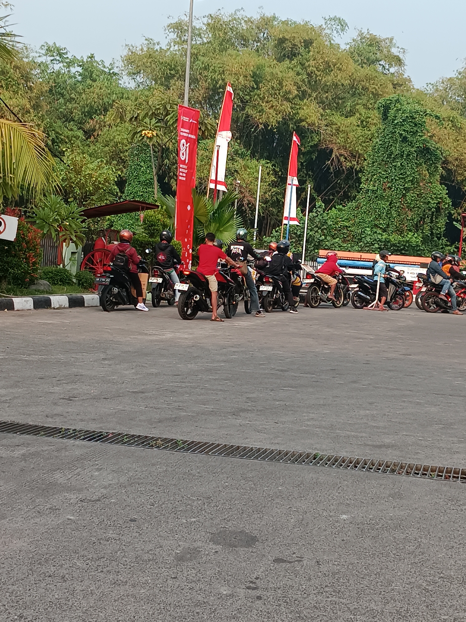 In Pleret, Indonesia, a group of people are gathered around motorcycles on a paved surface. The scene appears to be outdoors, with lush green trees forming a dense backdrop. Several red and white flags are prominently displayed, some bearing the number "81" in white text. The motorcycles are of various models, and several individuals are seated on them, suggesting a pause or gathering. The overall atmosphere is casual, with people dressed in everyday clothing. The pavement in the foreground features a metal grate, indicating it might be a drainage system. The time of day appears to be late afternoon, given the soft lighting and shadows.