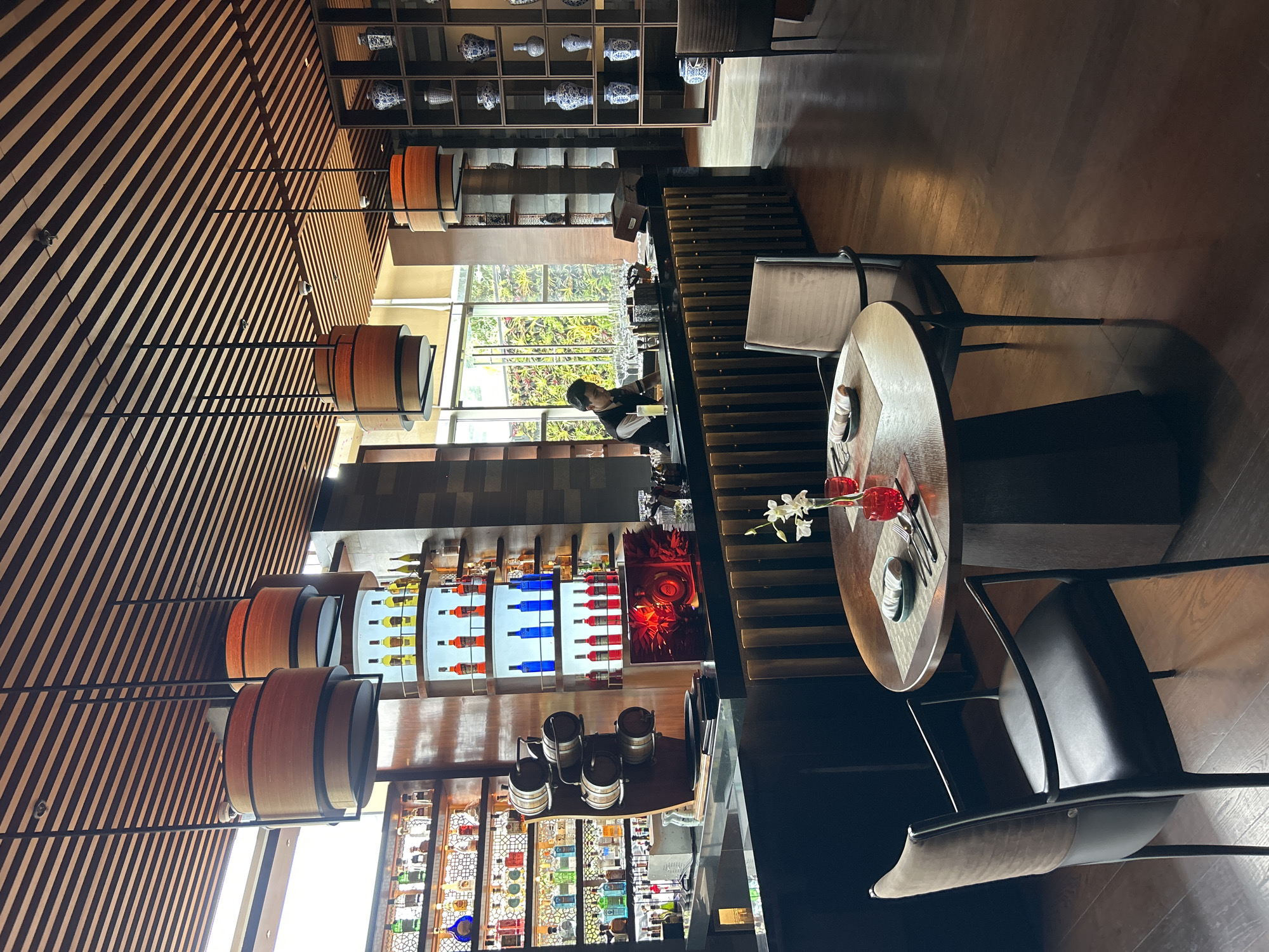 The image depicts the interior of a sophisticated bar and restaurant. The scene is set in Pune, India, at a location likely within a hotel or upscale establishment. In the foreground, a round wooden table is set for dining with plates, cutlery, and a small floral arrangement in a red vase. Two dark-colored chairs with upholstered seats and backs surround the table.
The bar area is prominent, featuring a long counter with a dark, reflective surface. Behind the counter, shelves are filled with an assortment of liquor bottles, some illuminated by colored lights. A bartender, wearing a dark vest over a white shirt, is visible behind the bar, engaged in preparing drinks. Large windows on the left offer a view of a lush green living wall, suggesting an outdoor or conservatory area.
Overhead, a distinctive ceiling design consists of parallel wooden slats. Several large, drum-shaped light fixtures with wooden exteriors hang from the ceiling, providing ambient illumination. To the right, a tall shelving unit displays decorative ceramic vases, adding to the refined aesthetic of the space. The flooring throughout the area is polished dark wood, reflecting the lighting and contributing to the overall luxurious ambiance. The restaurant appears to be either empty or in the early stages of operation, with no other patrons visible in the frame.