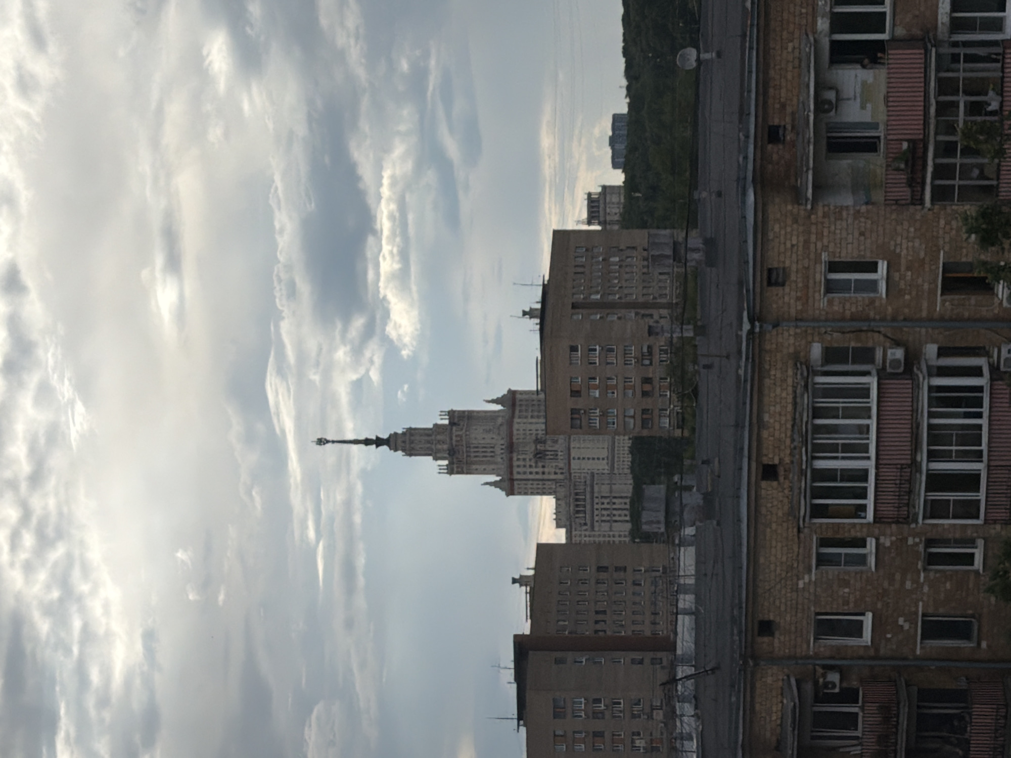 The image depicts a cityscape with a prominent tall, ornate building with a spire, likely a landmark, dominating the center. In the foreground, several multi-story apartment buildings with brick facades and balconies are visible. The sky is overcast with dramatic clouds. The overall scene suggests an urban environment under a cloudy sky.