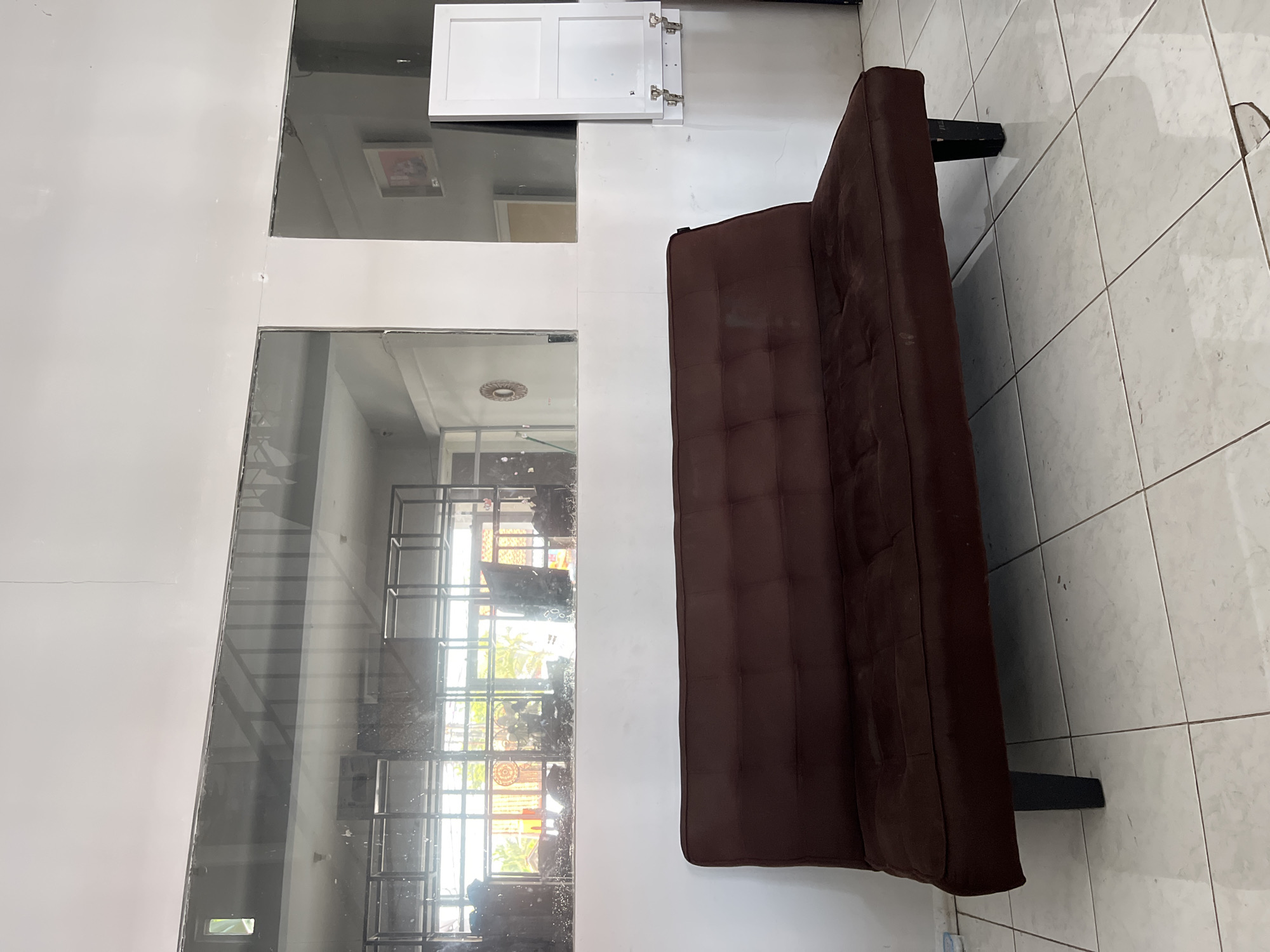 The image shows a dark brown futon sofa with tufted cushions, positioned centrally in the foreground on a tiled floor. The sofa has black legs and appears to be in a room with white walls. Above the sofa, a horizontal glass partition divides the space, revealing a reflection of a room with metal shelving units and some greenery outside. To the right of the futon, a white cabinet is mounted on the wall, with its doors partially open, revealing hinges. The overall environment appears to be an indoor space with a somewhat utilitarian aesthetic. There are no people or animals visible in the image. The location is identified as Kedawung, Indonesia.