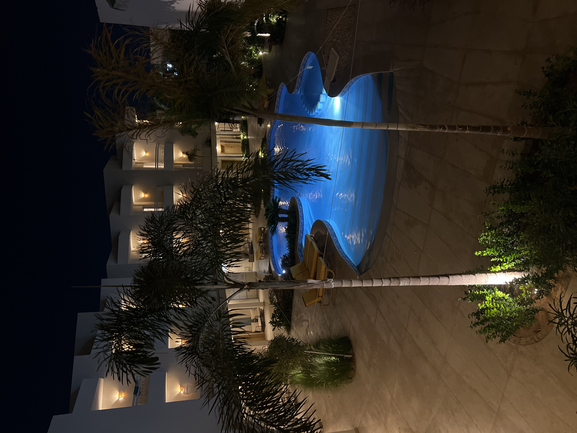 The image depicts a nighttime view of a resort pool area in Sharm El Sheikh, Egypt. A uniquely shaped swimming pool with bright blue illumination dominates the center of the scene. The pool area is surrounded by light-colored tiled paving. Several palm trees are visible, with their fronds casting shadows on the ground. The surrounding buildings are modern in design, with balconies and exterior lighting that casts a warm glow. Yellow lounge chairs are placed near the pool, suggesting a recreational setting. The sky is dark, indicating it is late at night. The scene is well-lit by artificial lights, creating a tranquil and inviting atmosphere.