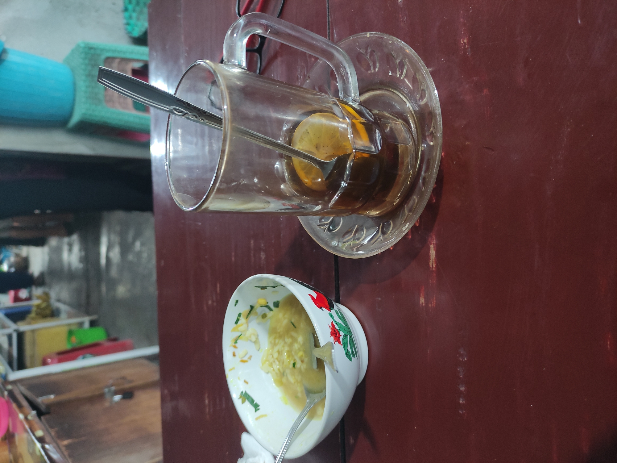 The image shows a close-up view of a dark wooden table, on which are placed a bowl of food and a glass of tea. The bowl, to the left of the frame, is white with a red rooster design and contains a light-colored, soupy dish with visible noodles and green garnishes. A spoon rests inside the bowl. To the right of the bowl is a clear glass mug filled with dark liquid, likely tea, and a slice of lemon. A spoon is also present in the mug. The glass sits on a clear glass saucer. In the blurred background, there appears to be an outdoor market or food stall setting with shelves, plastic containers, and indistinct figures of people. The location is identified as Gentan, Indonesia.
