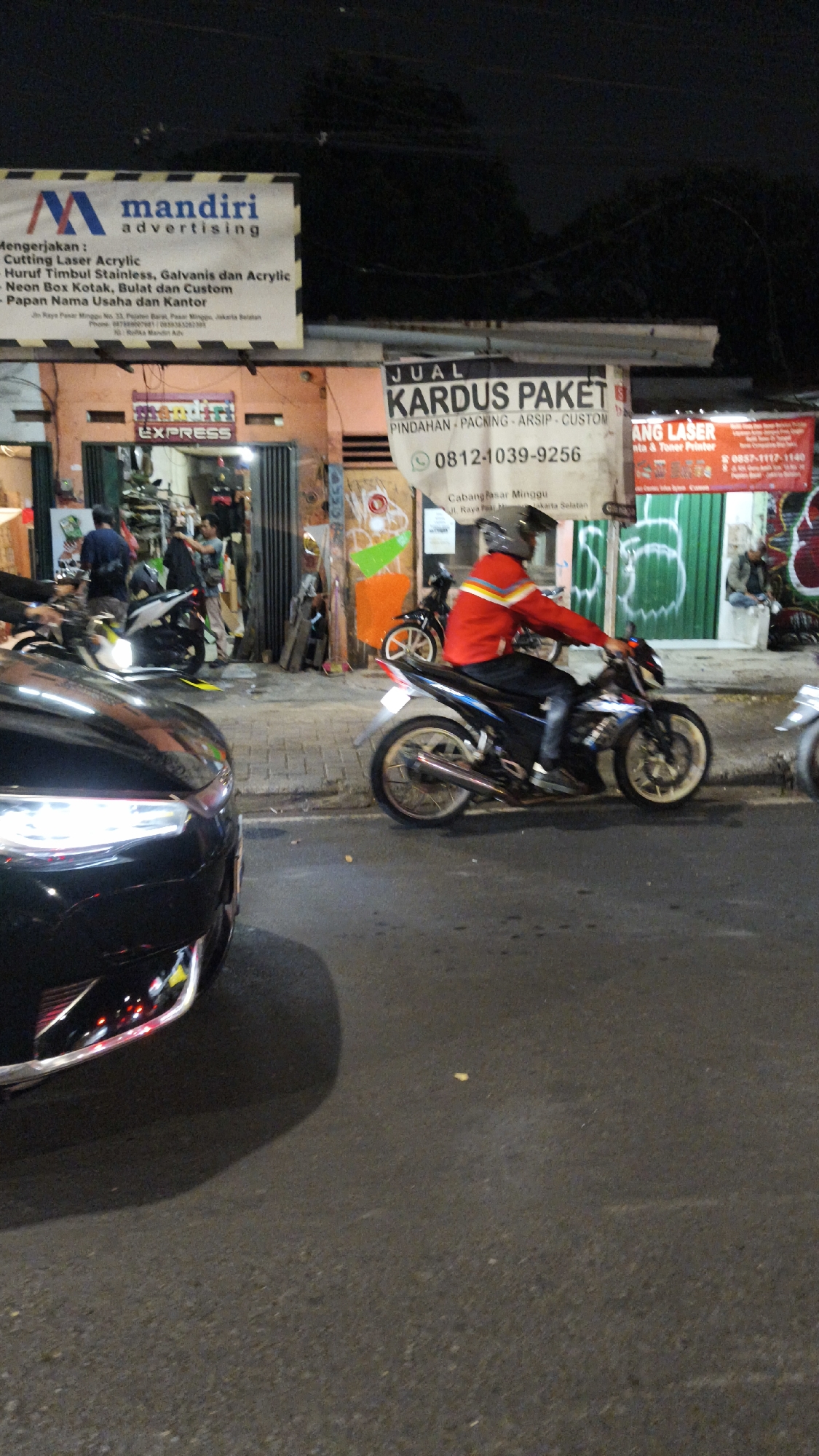 The image depicts a street scene at night in South Jakarta, Indonesia, likely an area with shops and businesses. In the foreground, a black car's front end is visible, reflecting lights from its headlights. To the right, a person on a motorcycle wearing a red jacket and a helmet is riding down the street. The motorcycle has a sleek design with silver accents. Behind the motorcycle, a shop entrance is visible with an orange and green facade. Signs are displayed above the shop, with one advertising "JUAL KARDUS PAKET" (Sale of Packet Boxes) and providing a phone number. Another sign above that, with the "M" logo, reads "mandiri advertising" and lists services like "Cutting Laser Acrylic," "Neon Box Kotak," and "Papan Nama Usaha dan Kantor" (Business and Office Nameplates). Further to the right, another shop displays a red banner with "ANG LASER" and what appears to be a phone number. The street is paved, and there are sidewalks with some debris and graffiti. The overall impression is that of a busy commercial area at night, with businesses operating and people on the move.
