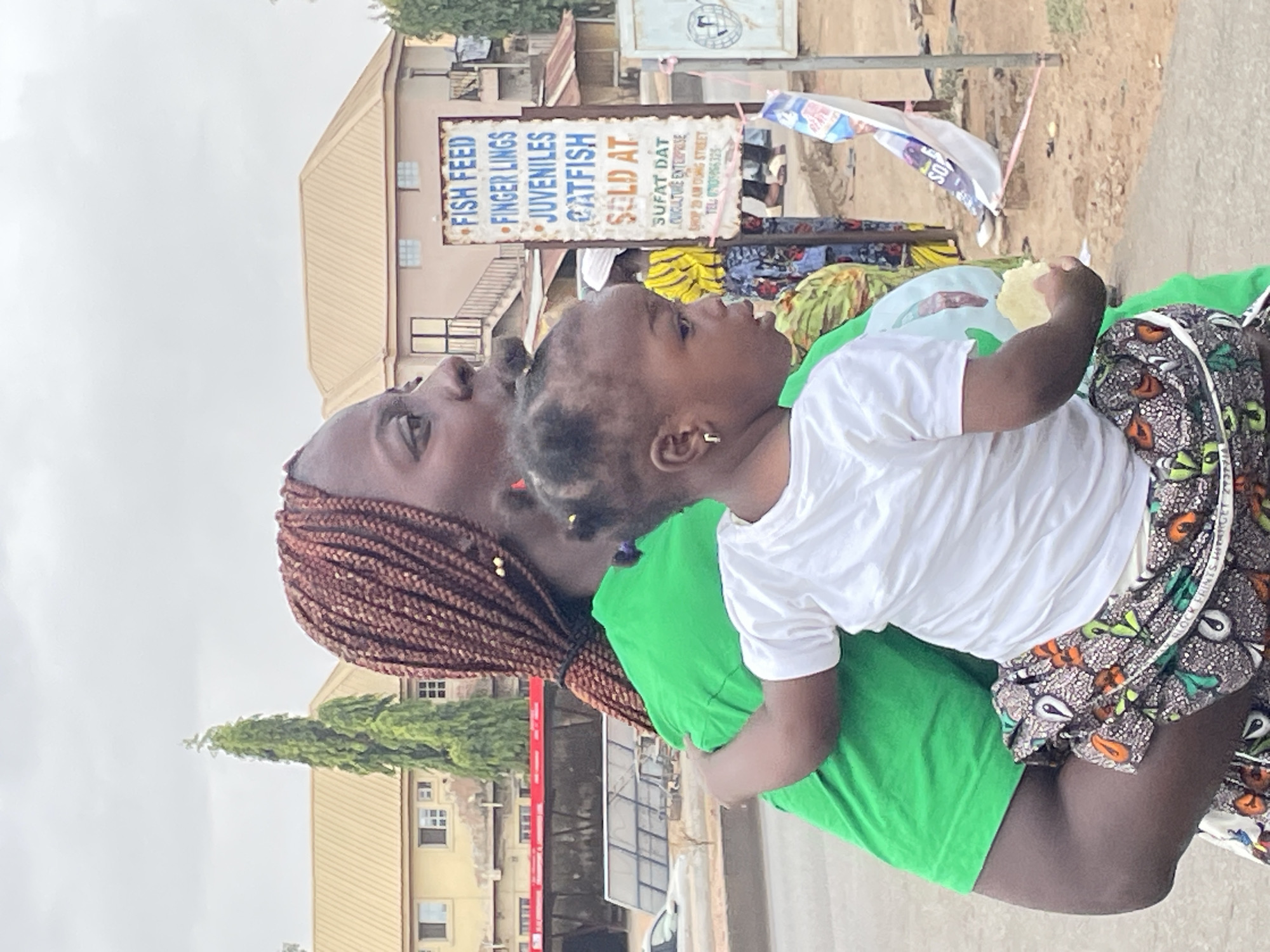 A woman with dark skin and brown braids holds a small child in her arms. The child has short, dark hair and is wearing a white shirt and patterned skirt. The woman is wearing a green shirt and a patterned wrapper around her waist.
The background includes buildings, trees, and a sign that reads "FISH FEED, FINGER LINGS, JUVENILES, CATFISH SOLD AT SUFAT DAT CUIculture ENTERPRISE SHOP 29 AM DUNG STREET TEL: 0705056325." This sign, and the shop address, indicate that the location is in Jos, Nigeria. The child is holding what looks like a piece of bread or food in their hand.