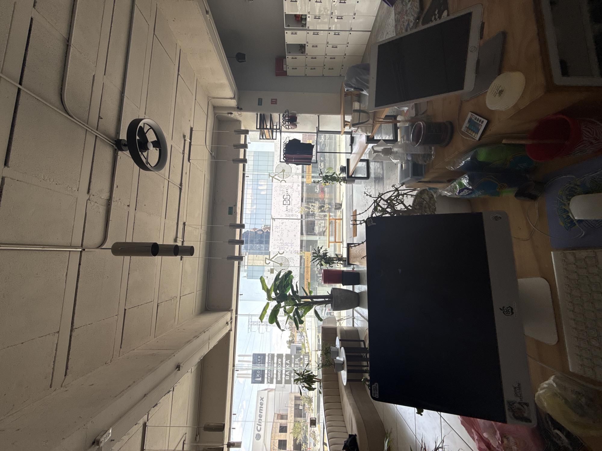 The image depicts the interior of a workspace, potentially an office, with a view overlooking a cityscape in Puebla, Mexico. The focus is on a desk area with two Apple iMac computers, a keyboard, and various desk accessories like a magic mouse, cups, plants, and stationery. The windows behind the desk show a partial view of the city with buildings bearing logos and names of businesses (e.g., "CINEMEX", "KOI FLEX", "CIUDAD MADERAS", "SWEATCORE", "BAJKA"). A rack with clothes is also visible. The interior also includes a series of lockers. Overhead, a ventilation system is visible along the ceiling and a circular fan is mounted on the wall. The overall impression is of a modern workspace with a prominent view of the surrounding urban area.