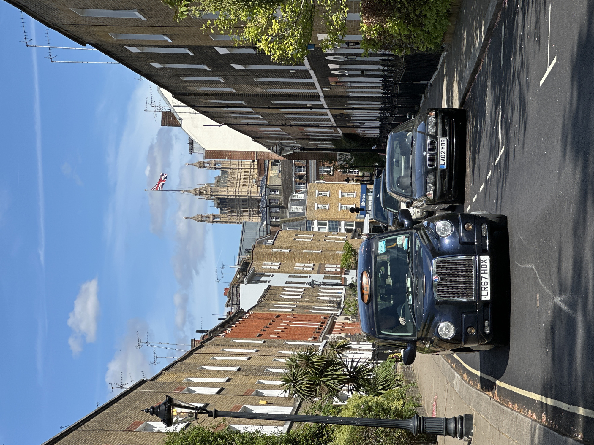 A street scene in London, United Kingdom shows brick buildings lining both sides of a narrow street. A black London taxi is in the foreground on the right side of the street. A dark-colored car is parked directly behind it. In the distance is the Palace of Westminster, with the Union Jack flag flying atop. The sky is blue with scattered clouds. A street lamp stands in the lower left corner.