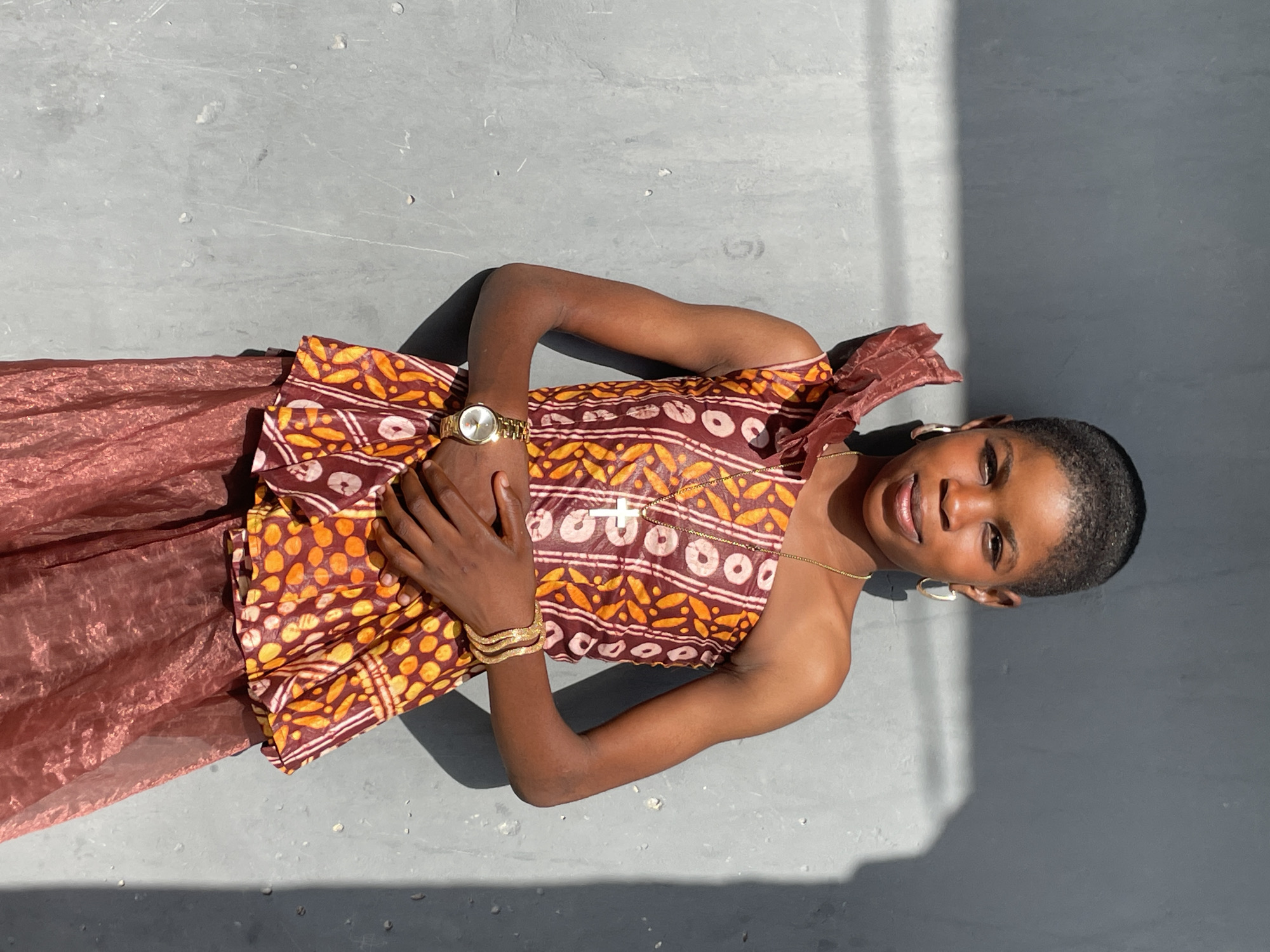 The image, captured in Badagry, Nigeria, presents a young person lying down on a light gray, textured concrete or paved surface. The subject has dark skin, very short dark hair, and is looking directly at the camera with a relaxed smile.
She is dressed in a sleeveless top featuring a vibrant, traditional-style pattern in shades of red-brown, orange, and yellow. A semi-sheer, reddish-brown skirt or fabric is visible below the top. Her arms are bent at the elbows, with hands clasped over her midsection.
Accessories include a gold-colored watch on her left wrist, multiple gold-colored bangles on her right wrist, a thin gold-colored necklace with a small white pendant (possibly a cross shape), and gold-colored hoop earrings.
The scene is illuminated by bright, direct sunlight, creating distinct, sharp shadows on the ground, suggesting it is likely midday or early afternoon. The background consists solely of the light gray surface, providing a simple, uncluttered setting. The overall impression is one of ease and warmth under the sun. There is no visible text in the image.