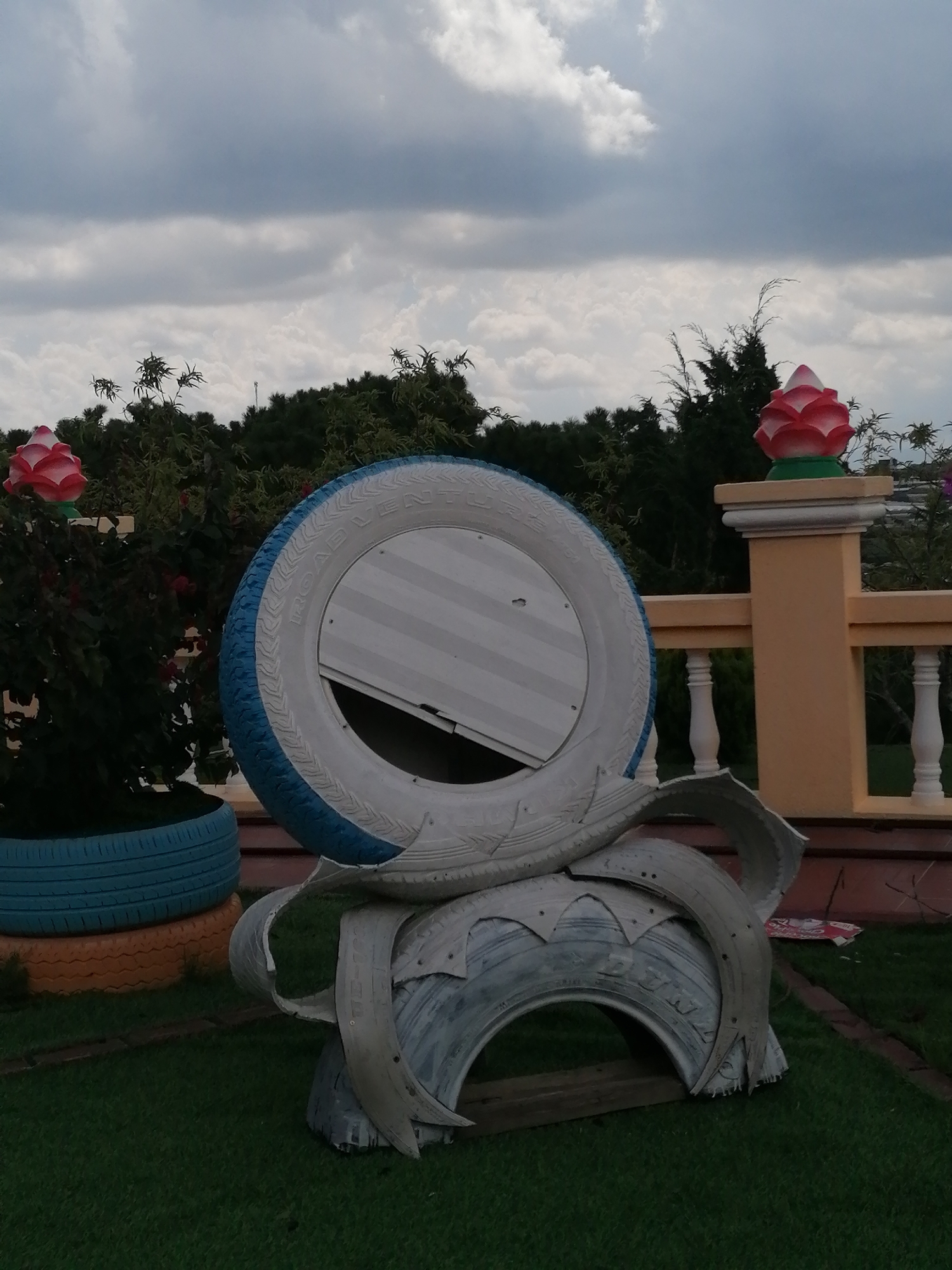 This image captures an outdoor scene in Lộc Quý, Vietnam, featuring a creative arrangement of repurposed tires. In the foreground, a large tire, painted white with a blue rim, is mounted on a structure also made from tires. The tire has a white, corrugated metal insert, and appears to be part of an art installation or seating. To the left, several stacked tires, one painted blue and another orange, form a base for plants. In the background, a fence with white balusters and a yellow pillar topped with a red and pink lotus-like ornament can be seen. Lush green trees and an overcast sky complete the environment. No people are visible in the frame.