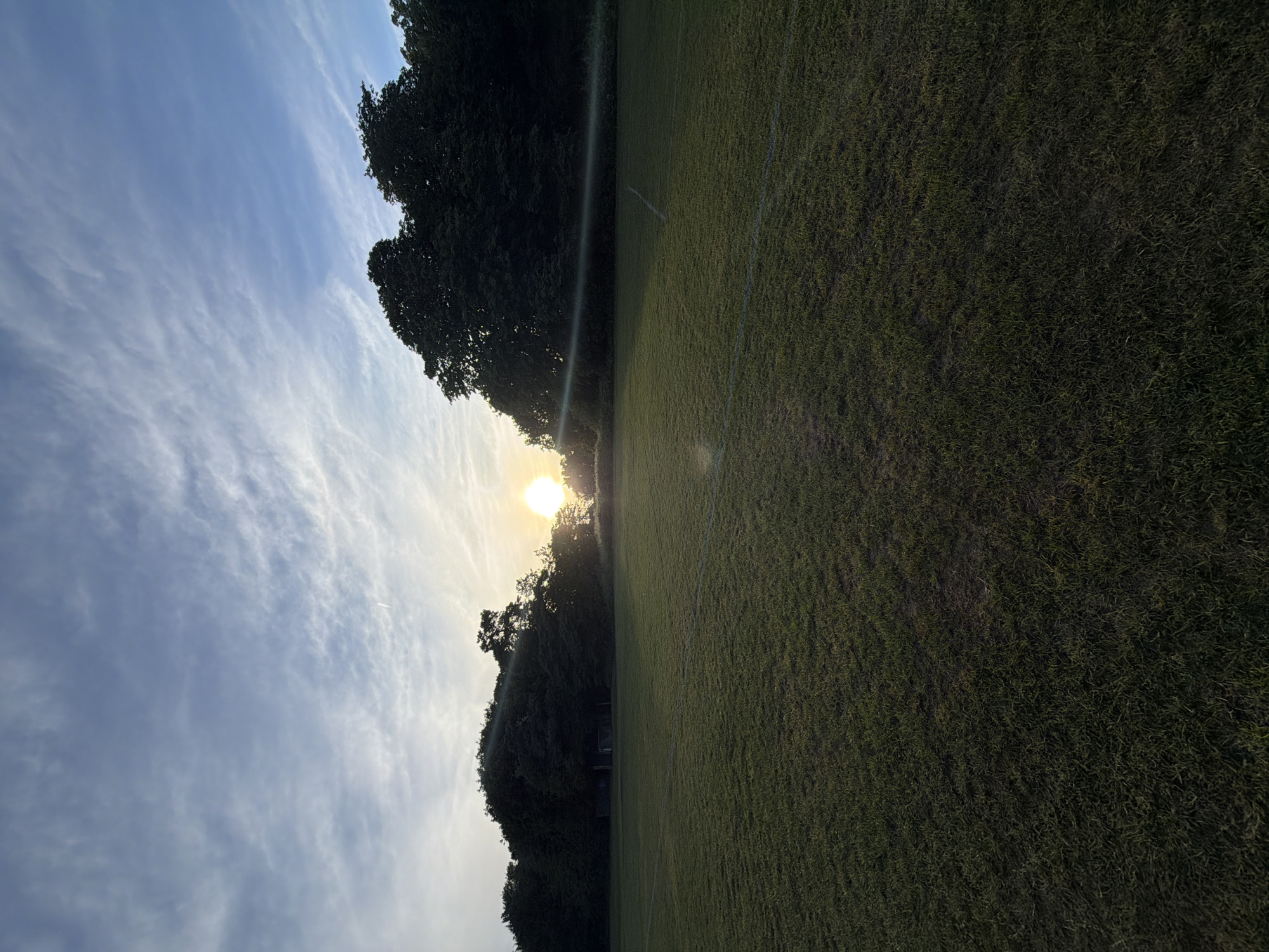 The image captures an expansive, dark green grass sports field under a golden hour sky, likely at sunset, in Maidstone, United Kingdom. White lines, typical of a football or soccer pitch, are visible on the grass, extending from the foreground towards the midground. A dense line of dark deciduous trees forms the background, appearing as silhouettes against the brighter sky and sun. A metal goalpost with netting is partially visible nestled within the trees on the left side of the frame. The bright, circular sun is positioned low on the horizon towards the right, partially obscured by the treeline, and casts a prominent lens flare across the image, along with long shadows extending across the field. The sky above is a gradient of light to deep blue, adorned with scattered, wispy white clouds. A faint airplane contrail is present in the upper right portion of the sky. The scene is devoid of any individuals, animals, or active human interactions.