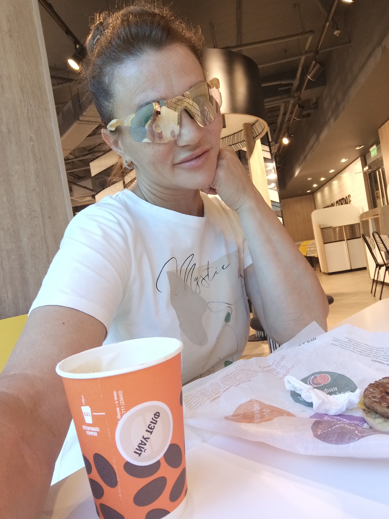 A woman with her hair pulled back in a bun, wearing large, reflective sunglasses and a white t-shirt with a graphic design, is taking a selfie at a table in what appears to be a fast-food restaurant. She is resting her chin on her hand and looking at the camera. In front of her on the table is an orange paper cup with a dark oval pattern, partially filled with a light-colored beverage, and a white wrapper with food items, including a burger. The background shows the interior of a restaurant, with booths, tables, chairs, and lighting fixtures visible. The lighting is bright, with overhead lights and some natural light possibly coming from a window or entrance. The overall environment suggests a casual dining setting, likely in Sochi, Russia, given the Cyrillic text on the cup and wrapper.