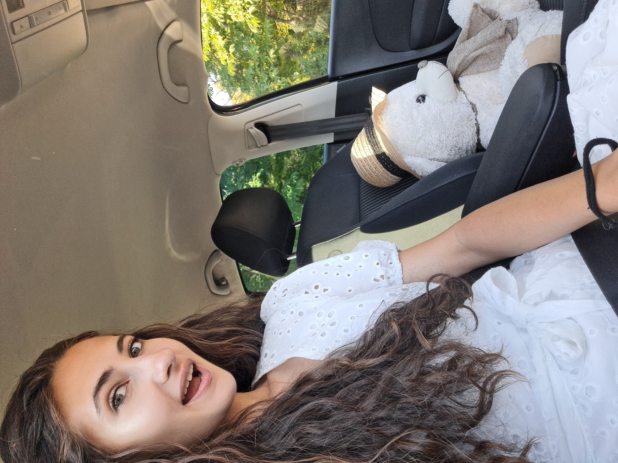 A young woman with long, dark, curly hair is in the passenger seat of a car, looking at the camera with an open mouth and surprised expression. She is wearing a white eyelet top with puffed sleeves. To her right, a white teddy bear sits on the car seat, wearing a straw hat and a light brown scarf. Outside the car window, green trees are visible, suggesting an outdoor or suburban setting. The interior of the car shows a gray headliner, a grab handle, and a black seatbelt. The image is taken from a slightly low angle. There are no specific landmarks visible that would indicate Sofia, Bulgaria.