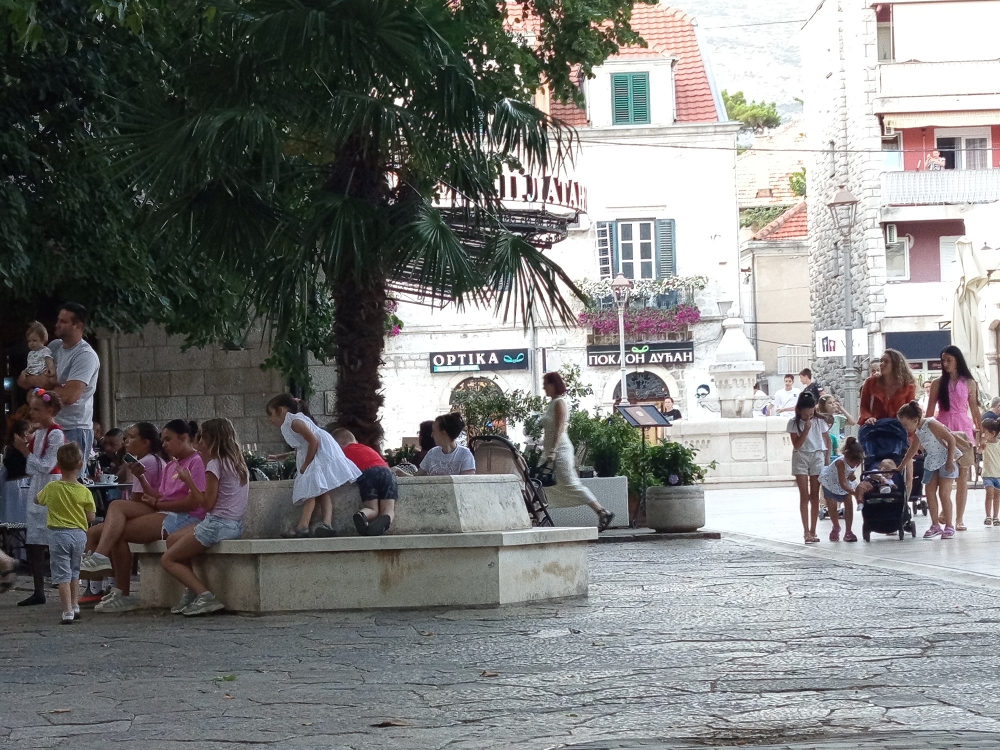 In Trebinje, Bosnia and Herzegovina, an outdoor scene depicts a plaza with several people, primarily children, engaged in various activities. A stone bench serves as a focal point, with a group of girls sitting on it. One girl in a white dress stands near the bench, looking towards a stroller. A man is holding a baby. The background features buildings with traditional architecture and greenery. The ground is paved with cobblestones. The overall atmosphere suggests a public gathering or leisurely outing.
