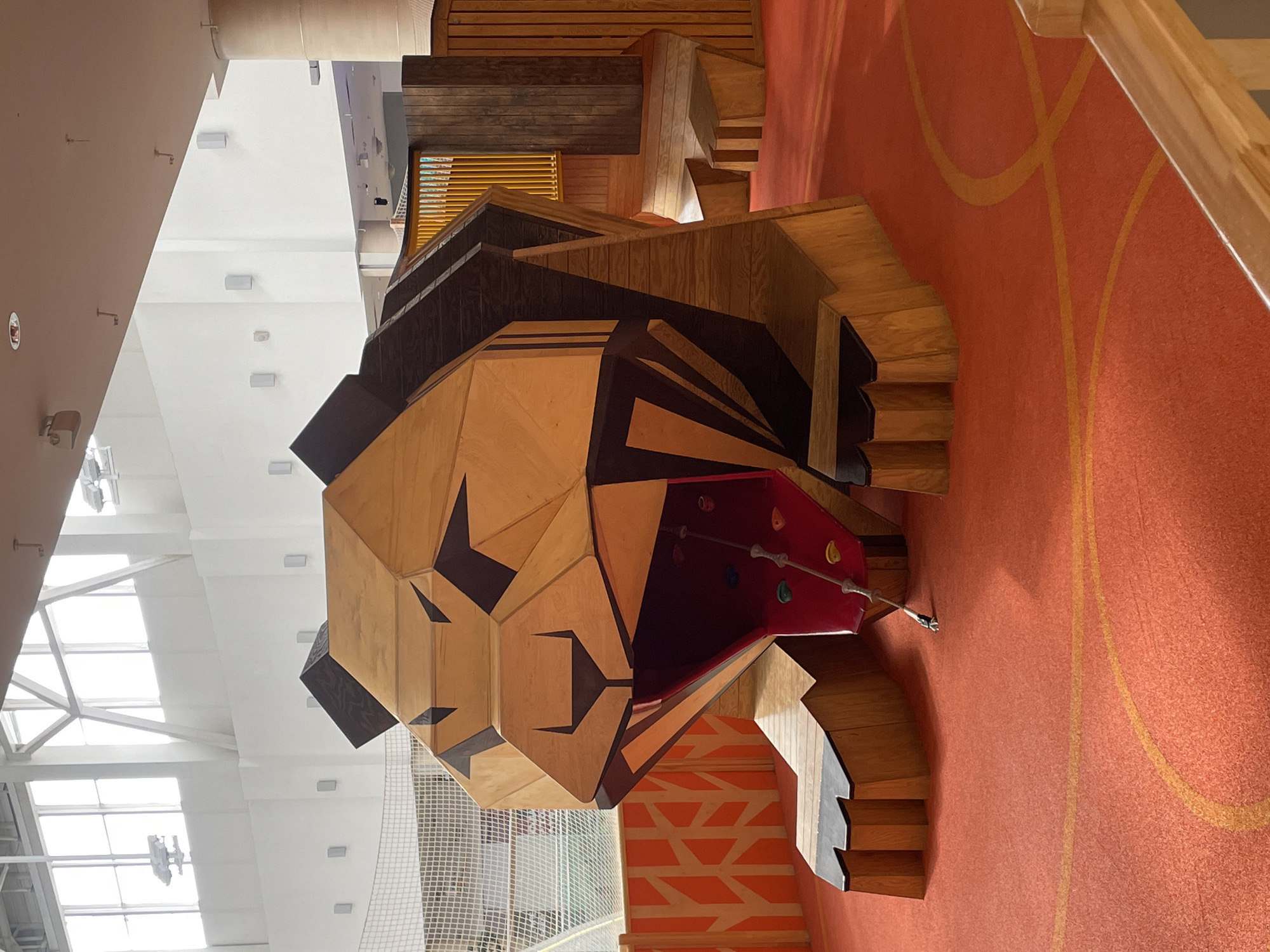 The image shows a large, polygonal wooden structure resembling a lion's head inside what appears to be an indoor playground. The head is predominantly light brown with dark brown accents defining the features. A red climbing surface with colorful handholds extends from the lion's mouth. The structure is built on a red floor with a yellow linear design. In the background, there's a white wall with large windows and a net. The location is Krasnoyarsk, Russia, as indicated by the source.